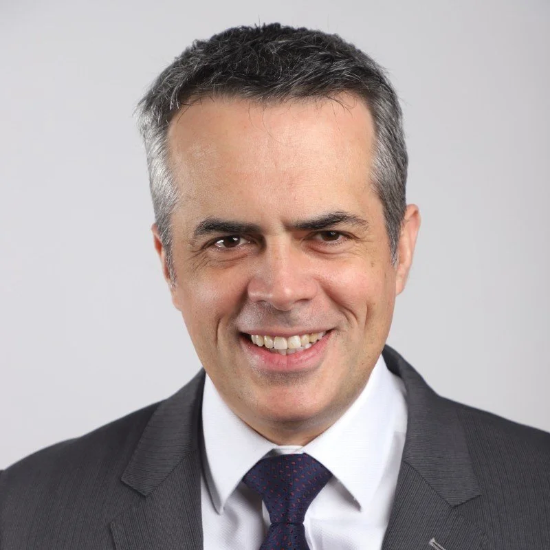 A professional headshot of a man with dark and gray hair, wearing a dark suit, white shirt, and dark tie, smiling at the camera against a plain light background.