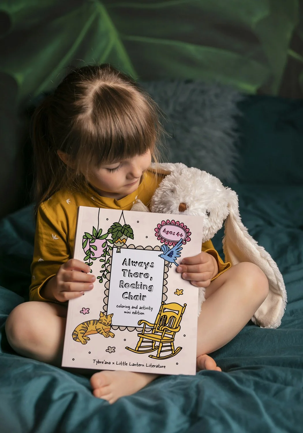 A young girl with brown hair and a yellow shirt sitting cross-legged on a blue bed, holding an illustrated coloring activity book titled 'Always There, Rocking Chair' with a plush bunny toy hugging her.