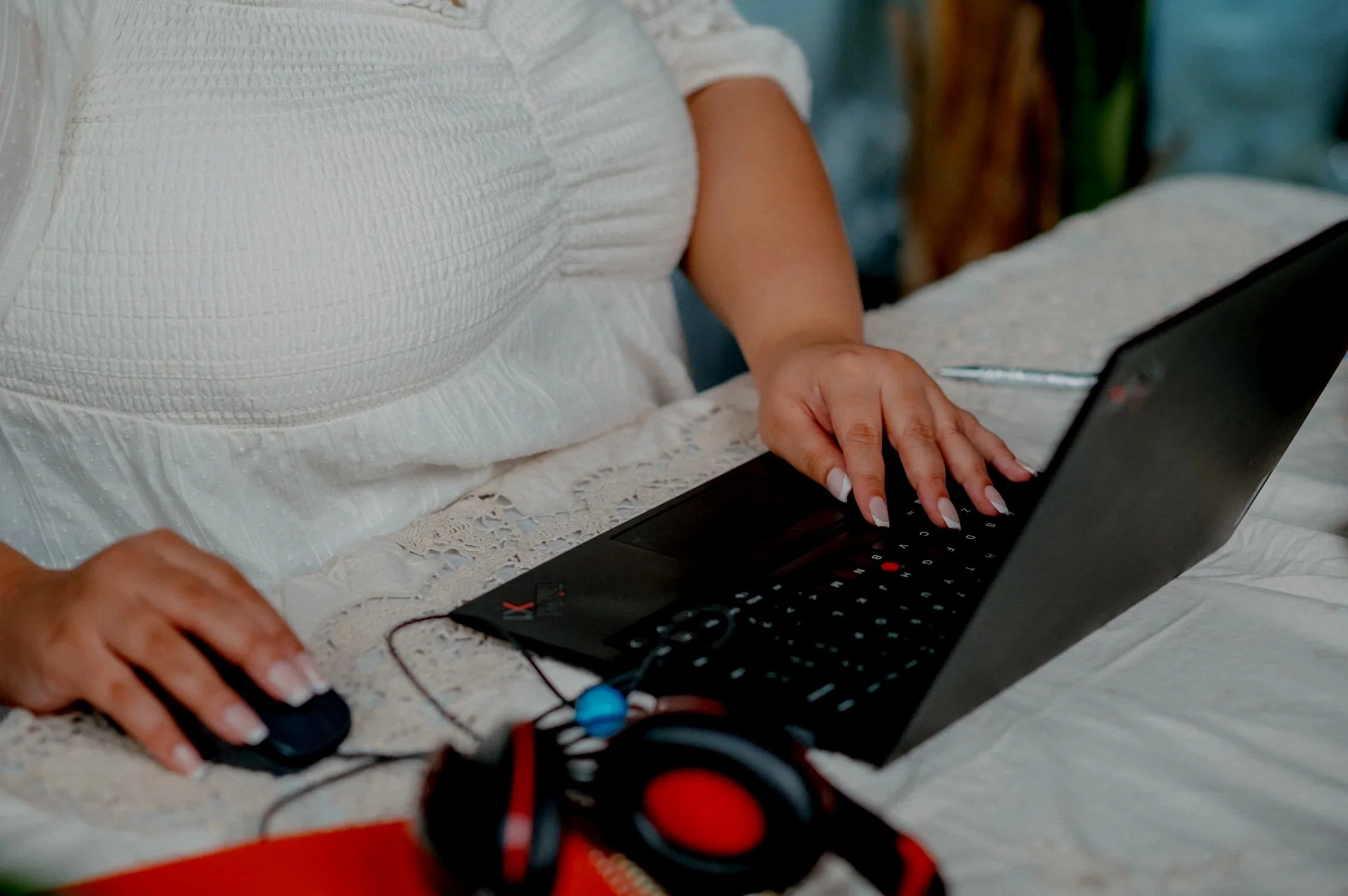 Person in white blouse using a black laptop with headphones and a wired mouse on a bed.