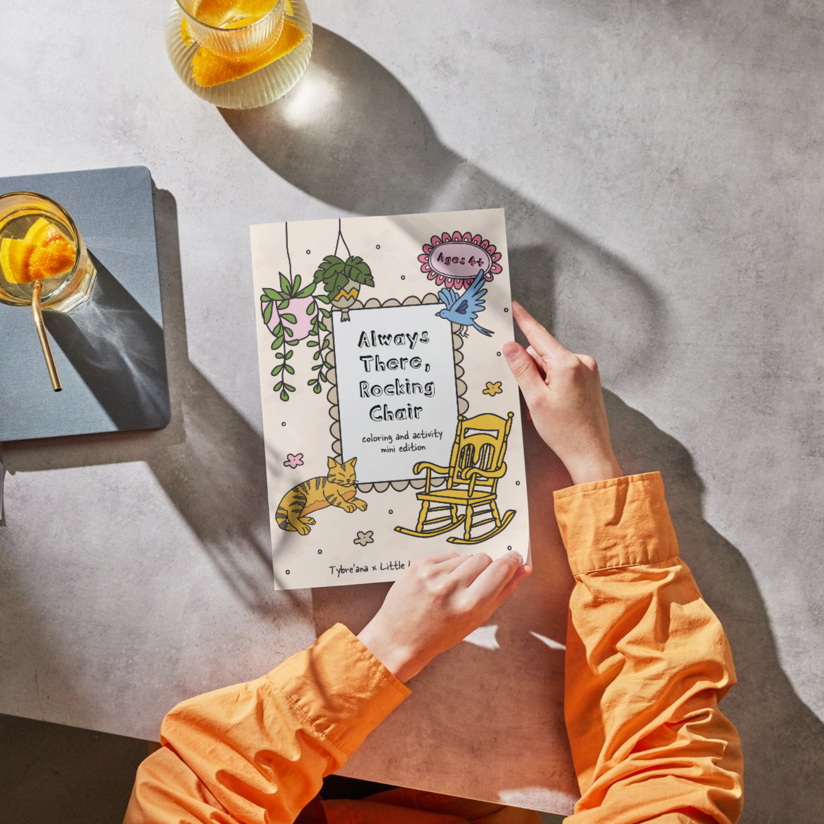 Child with orange sleeves holding a coloring book titled 'Always There, Rocking Chair' on a gray table with two glasses of lemonade with lemon slices and straws nearby.