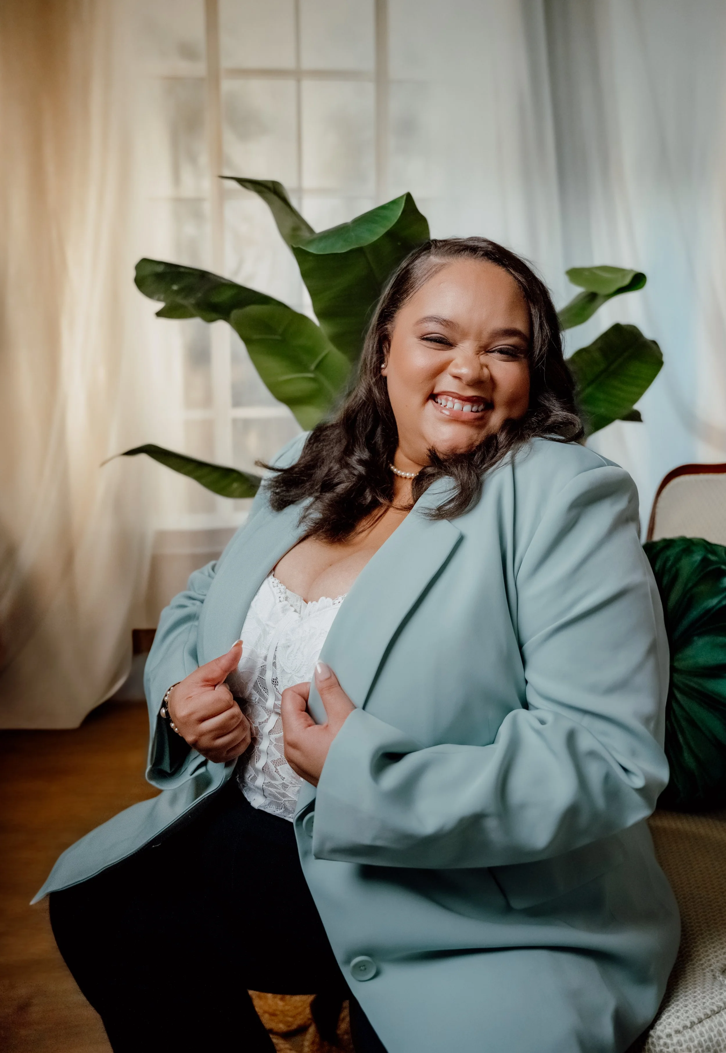 A joyful woman with dark hair in loose curls, wearing a light blue blazer over a white lace top, smiling and showing her teeth, standing in a bright room with large green plants and soft curtains.