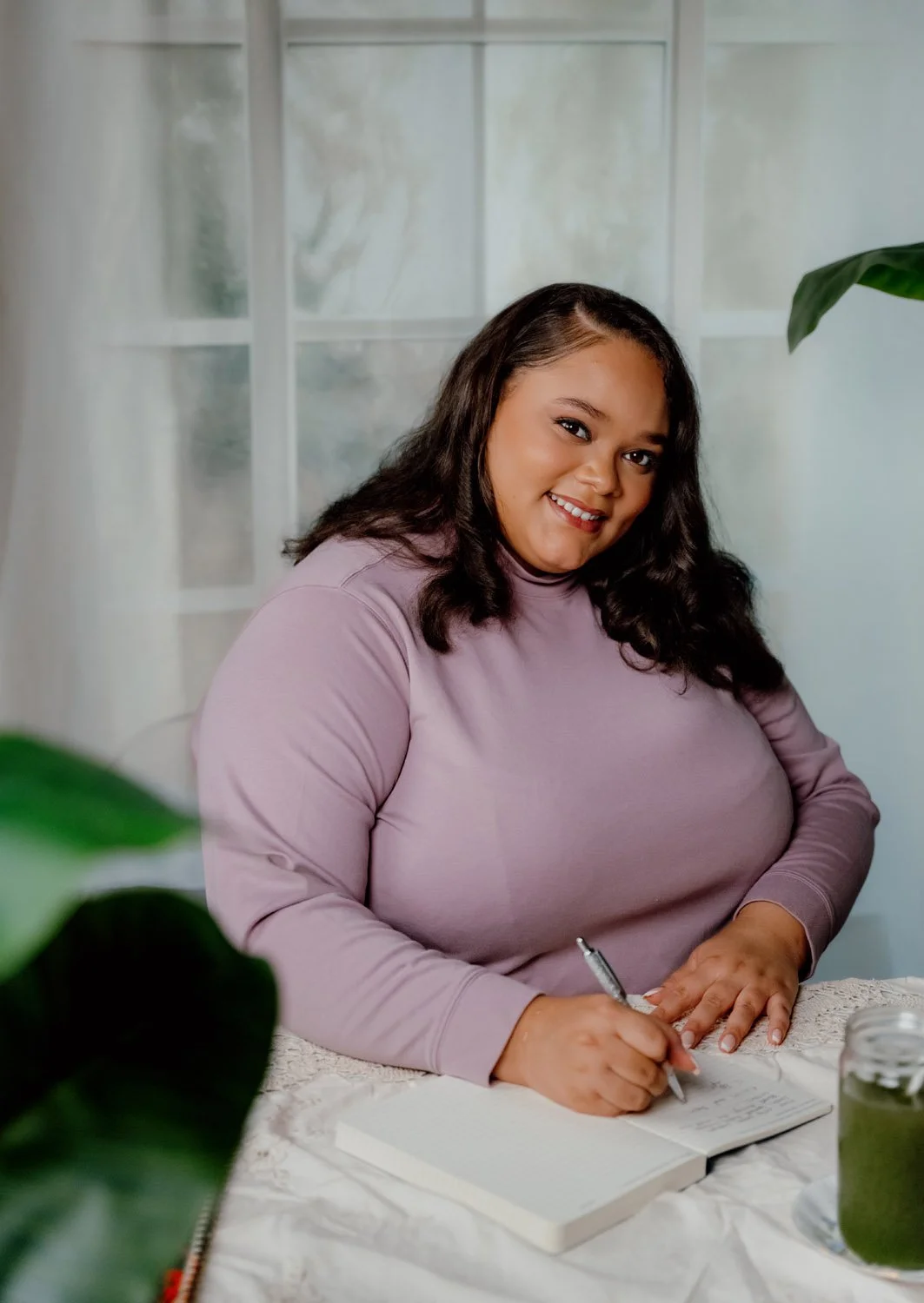 A woman with dark, wavy hair smiling and writing in a notebook at a table with a jar of green smoothie and a green plant in the foreground.