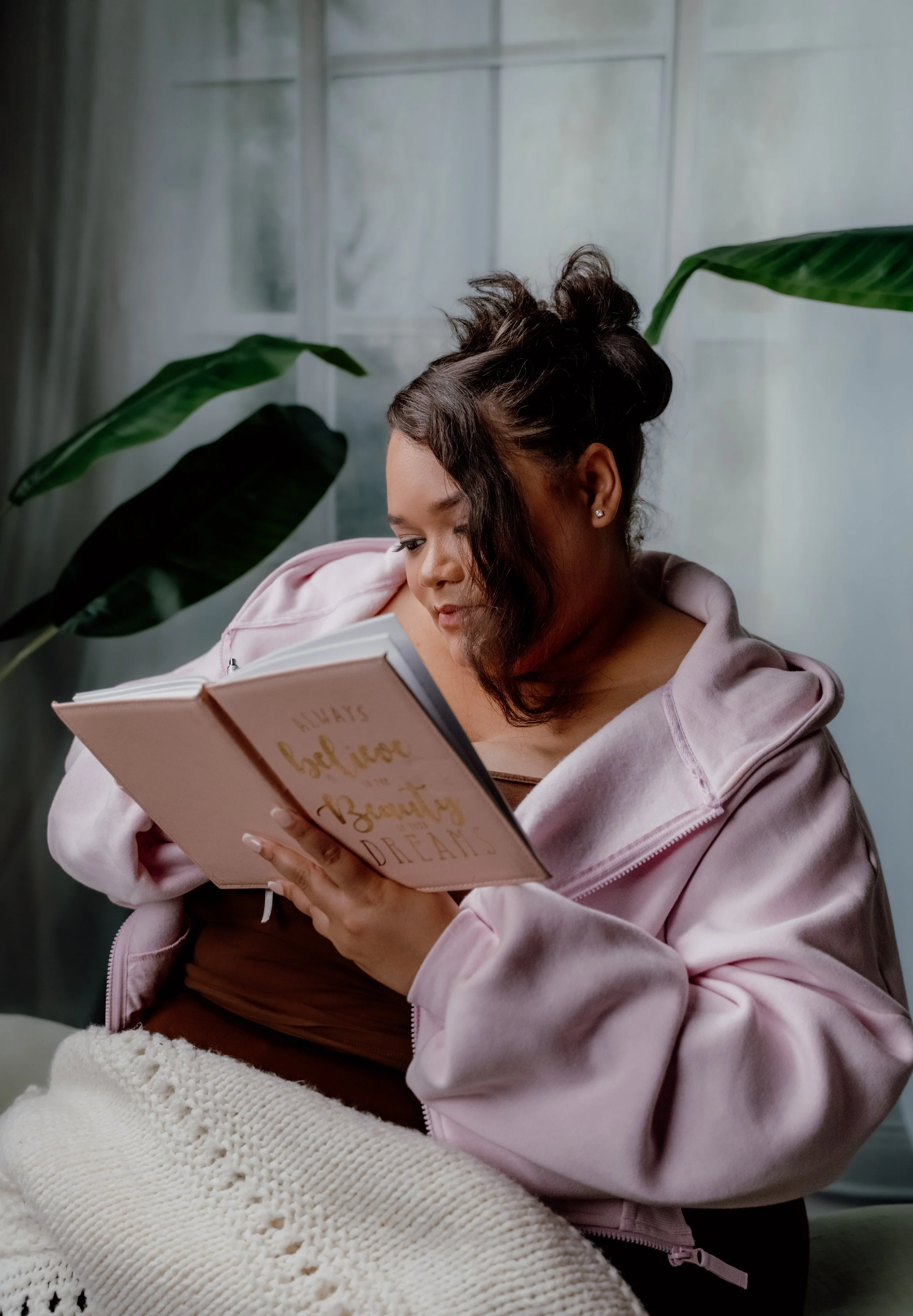 A woman with dark hair styled in a messy bun, wearing a pink hoodie, is sitting and reading a book titled "Always believe in yourself and your dreams." She is surrounded by green indoor plants in a softly lit room.