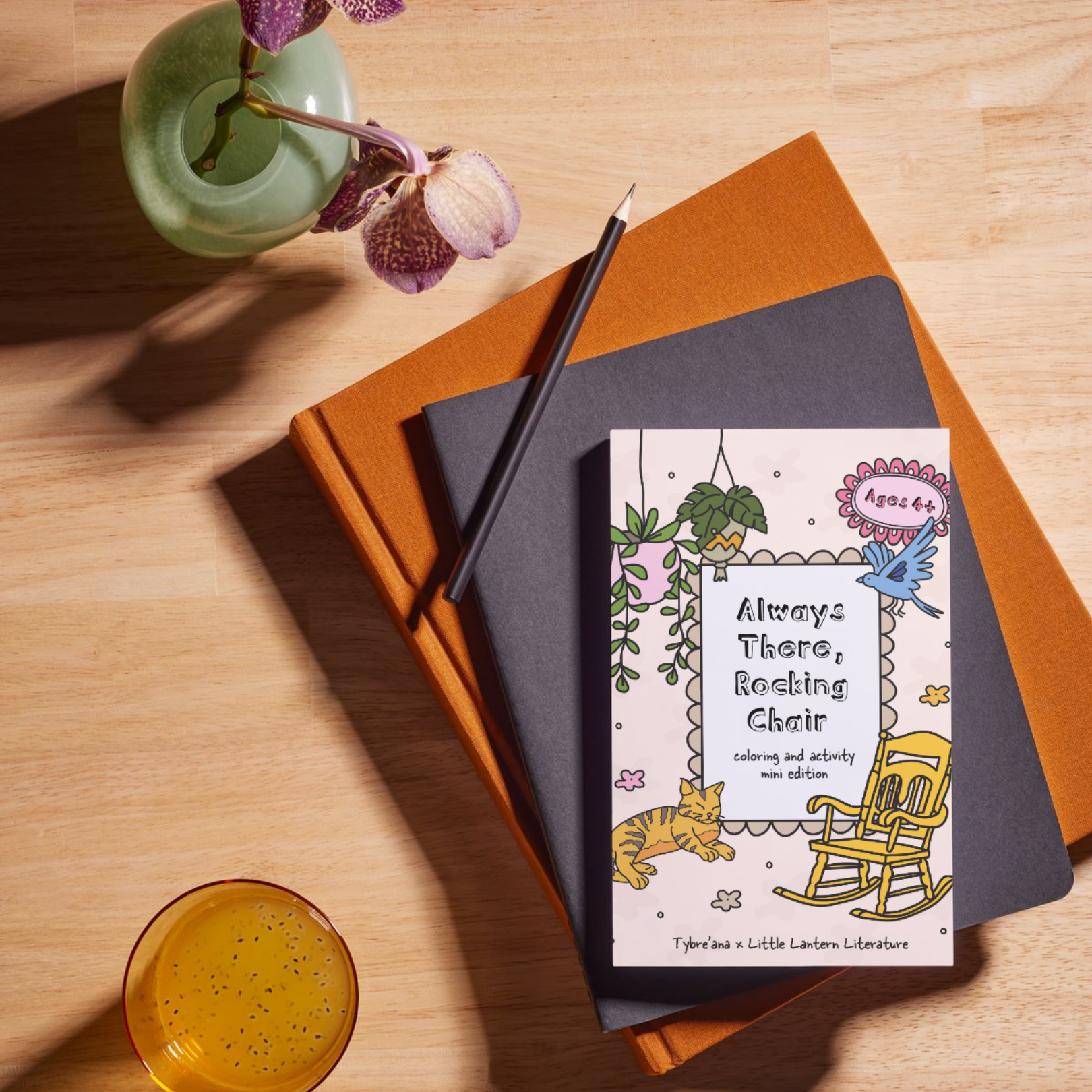 A stack of three books with a coloring book on top titled 'Always There, Rocking Chair' featuring a drawing of a rocking chair, a cat, a bird, hanging plants, and a sticker that says 'Ages 4+' on a wooden table. There is a pot with purple orchids, a black pencil resting on the books, and a glass of yellow liquid nearby.