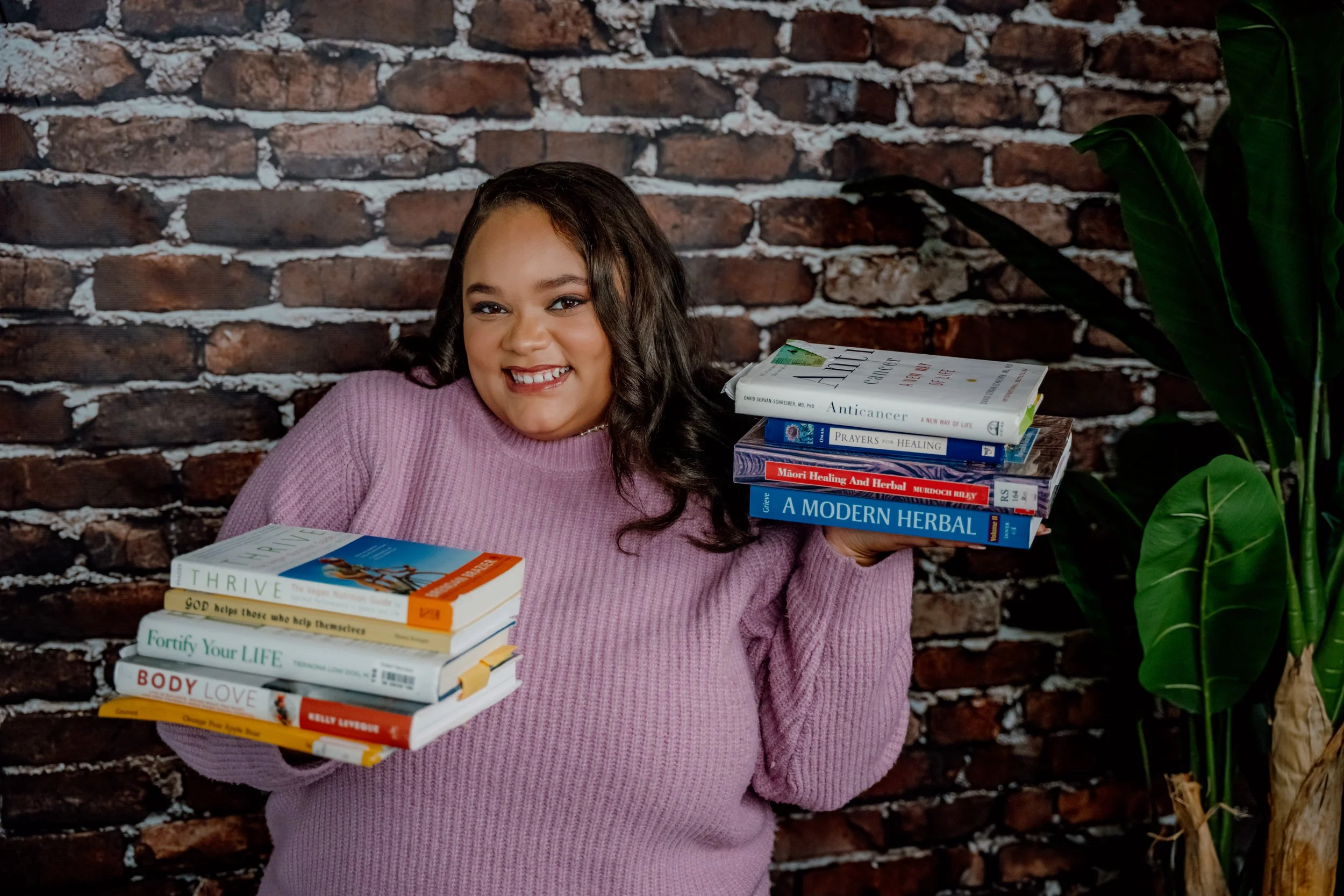 A young woman with long wavy dark hair smiling, wearing a pink sweater, standing in front of a brick wall, holding multiple books about health and wellness.
