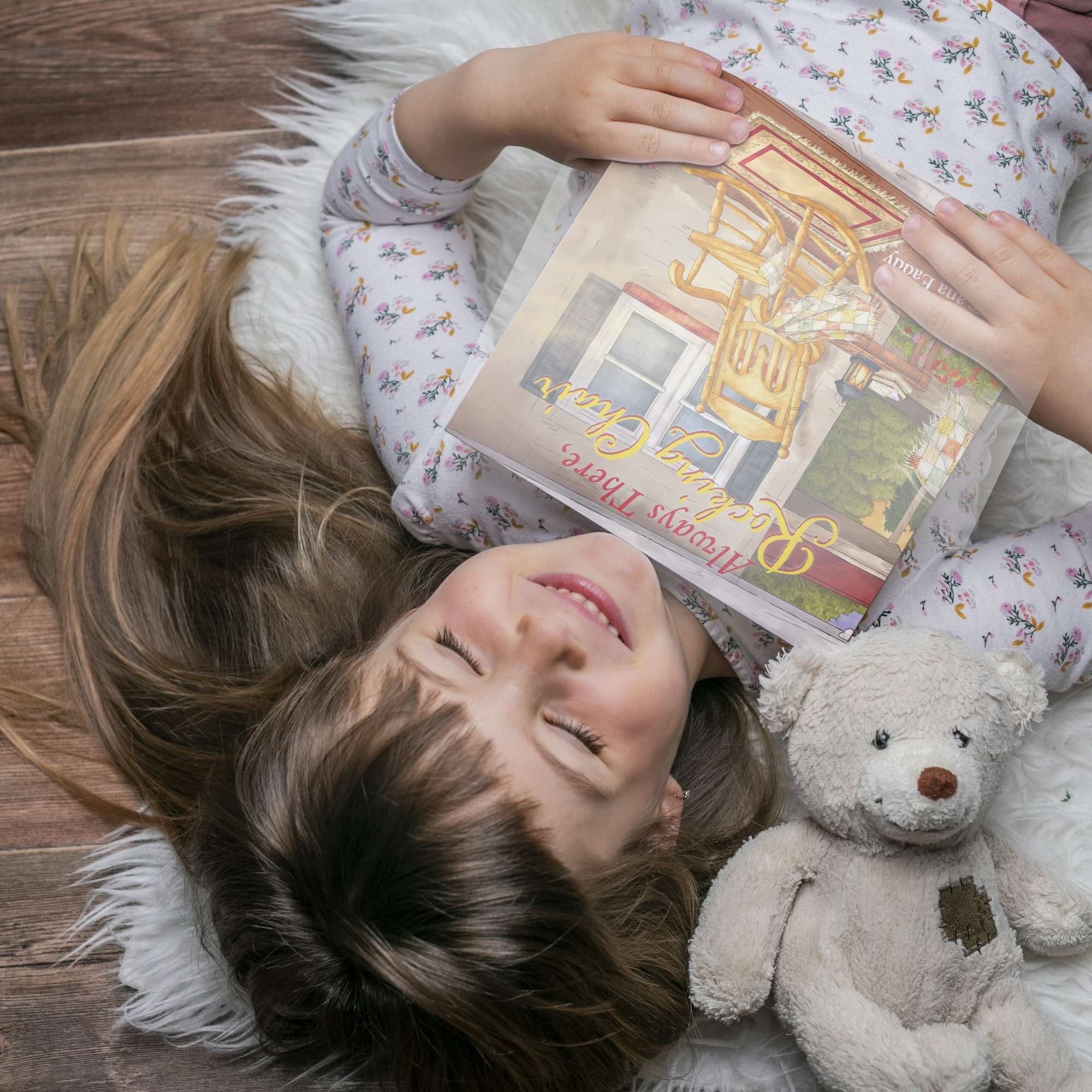A young girl with long brown hair is lying on a fluffy white rug on a wooden floor, smiling with her eyes closed. She is holding a children's book titled 'Mumsy TBexie & the C...' upside down. A worn teddy bear with a brown nose and a patch on its chest is beside her.
