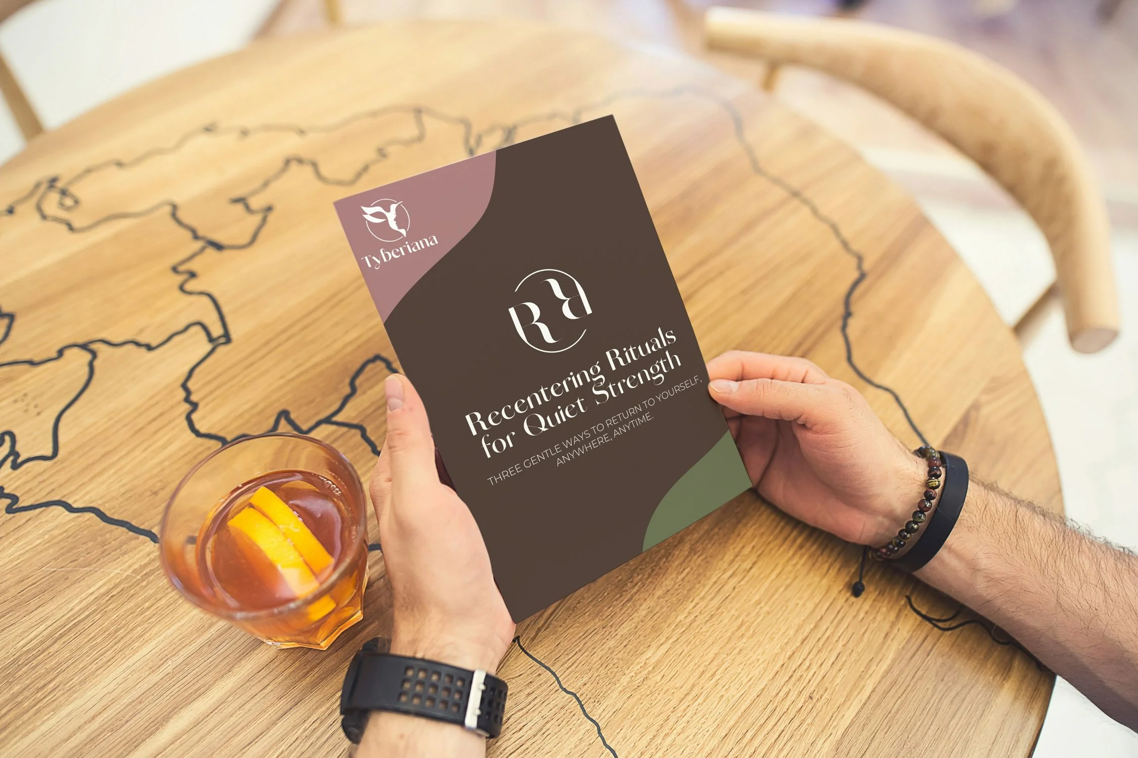 Person holding a pamphlet titled 'R4 Centering Rituals for Quiet Strength' with a glass of iced tea with lemon slices on a wooden table with a map outline