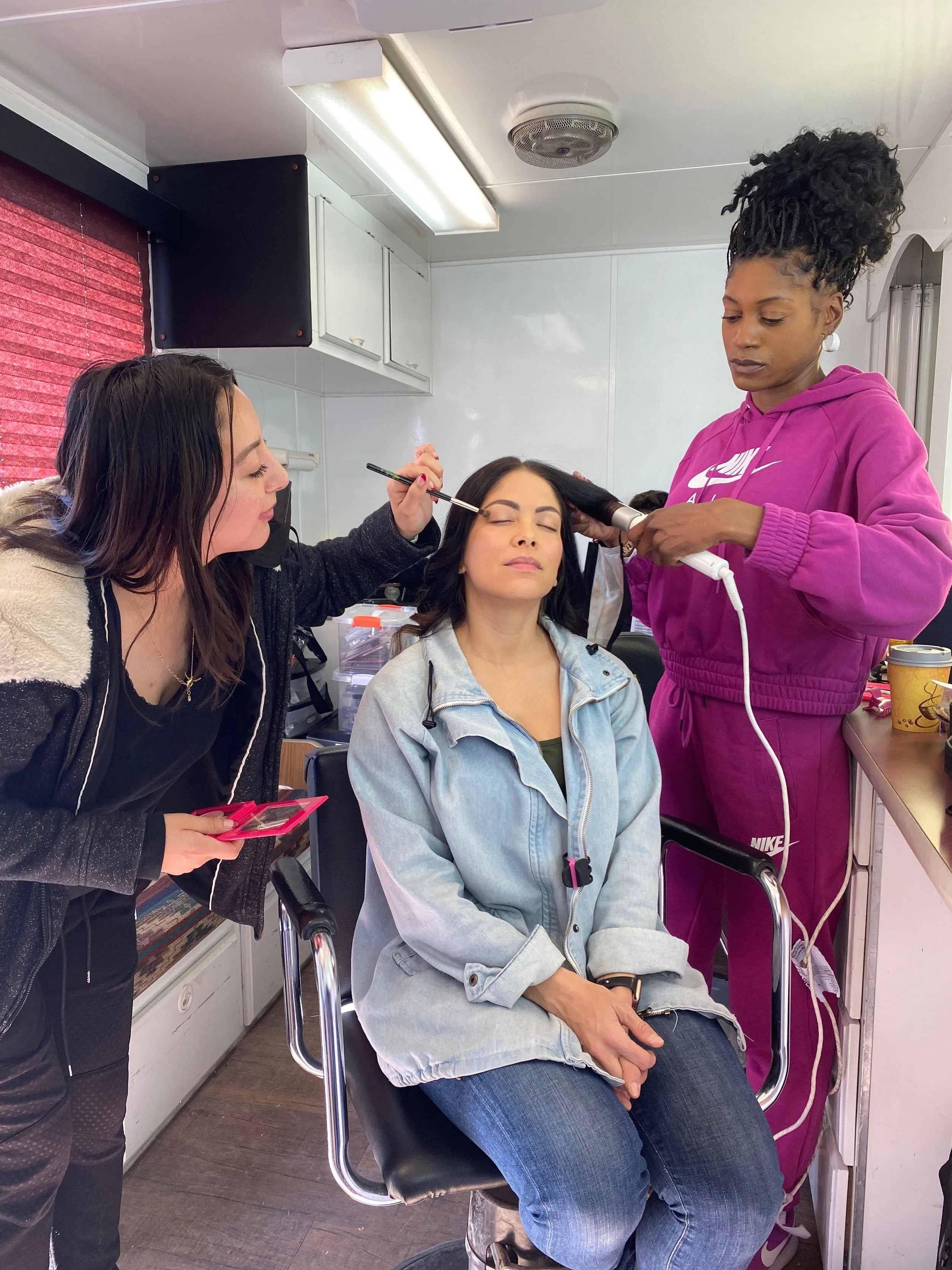 A woman seated in a salon chair getting her hair and makeup done, with a makeup artist applying eyeshadow and another stylist using a curling iron on her hair.