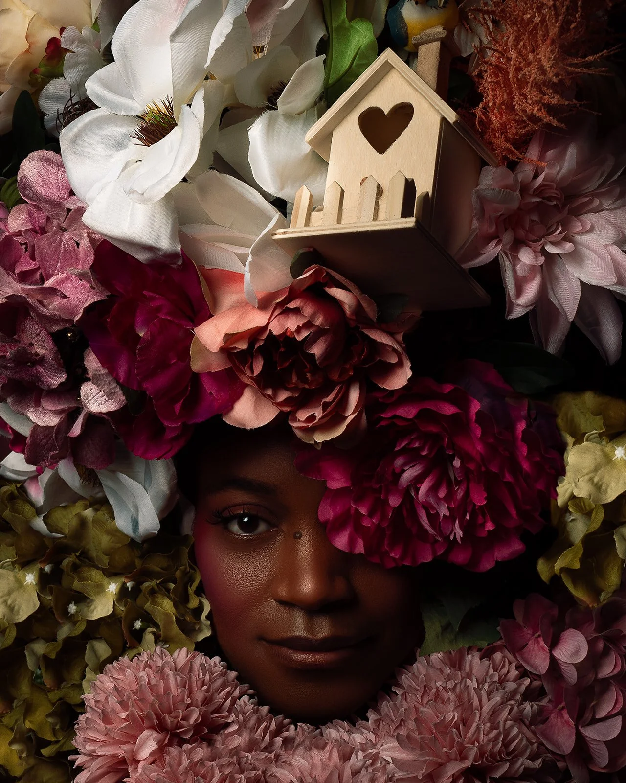 A woman with dark skin surrounded by and partially covered by a variety of colorful flowers, with a small wooden house with a heart-shaped cutout resting among the flowers on her head.
