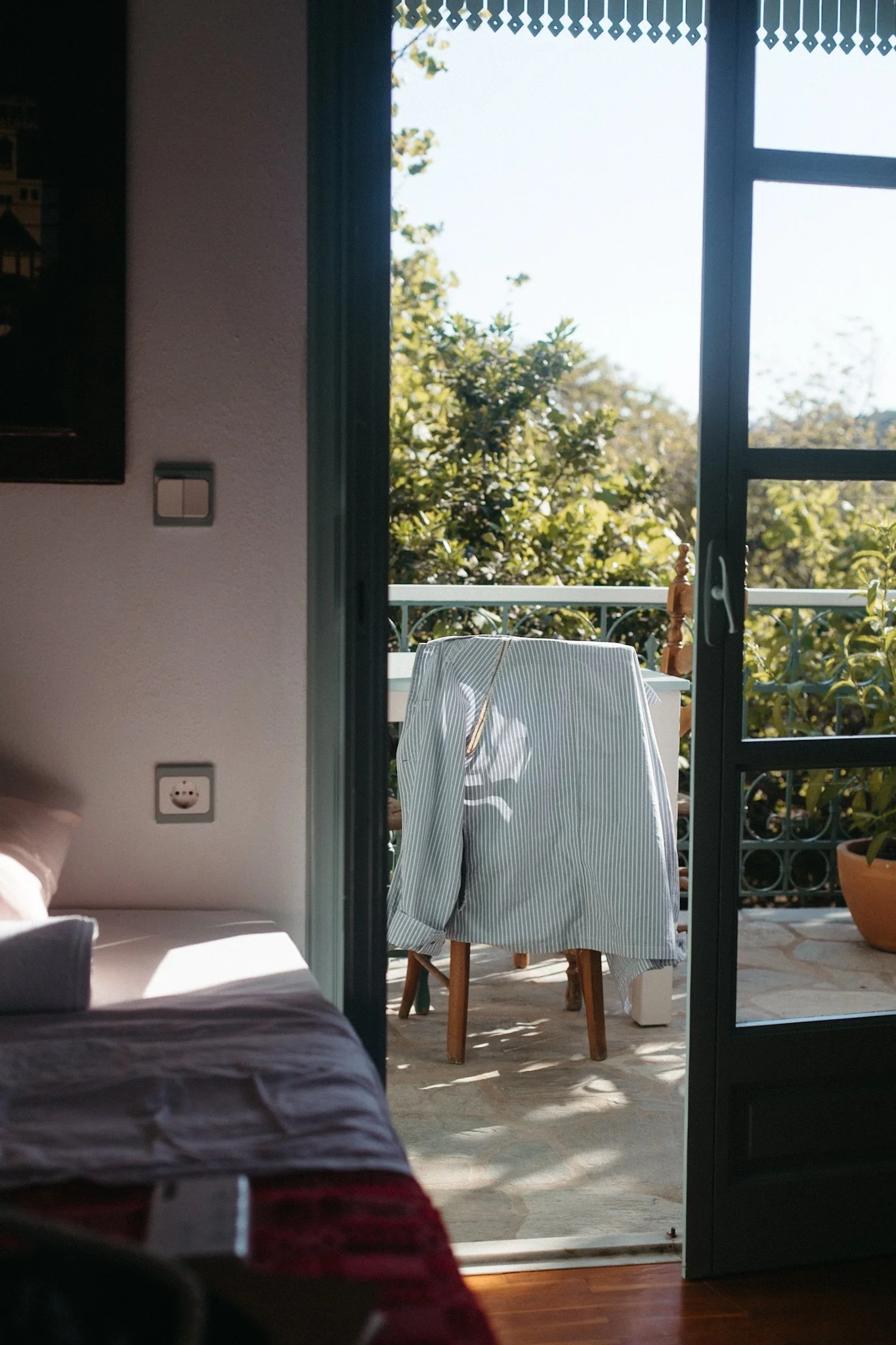 A simple bedroom with two single beds, a small wooden desk with a chair, and a balcony with outdoor chairs. There are paintings on the wall, a mirror above the desk, and windows letting in natural light. The room has light-colored walls and wooden flooring.