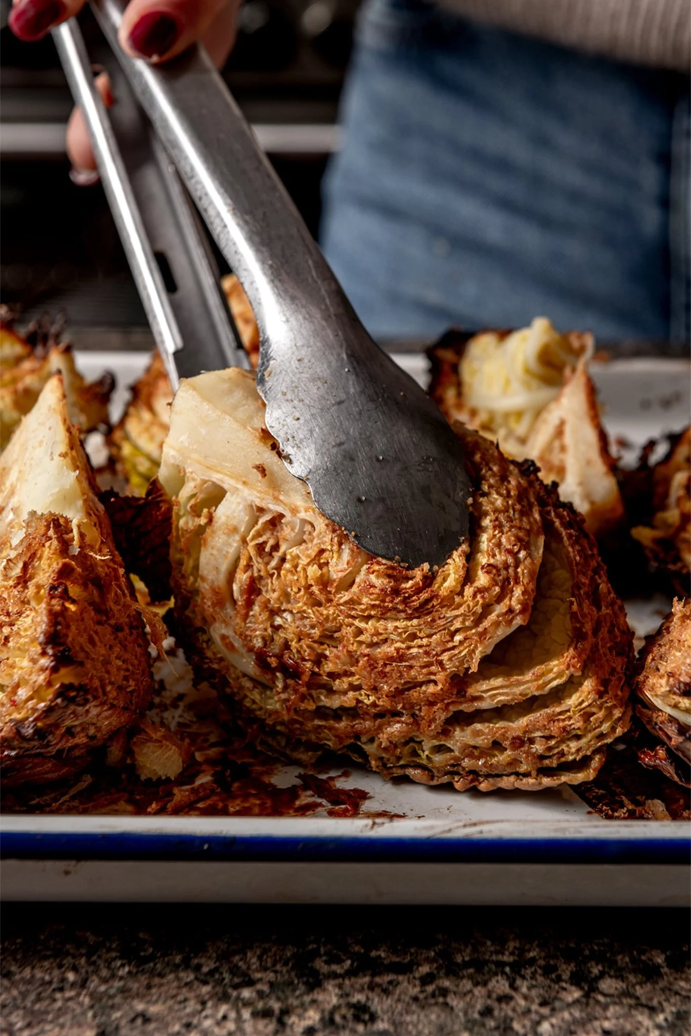 Hands using tongs to turn caramelised Savoy cabbage wedges on a baking tray mid-roast, with the sauce darkened and crisped against the leaves.