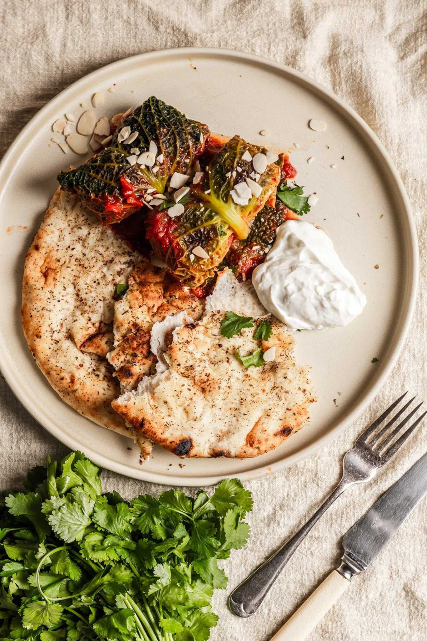 A plate with two lamb and apricot stuffed cabbage rolls in harissa tomato sauce, served alongside a warm flatbread, a spoonful of plain yoghurt, and scattered with toasted almonds and fresh coriander.