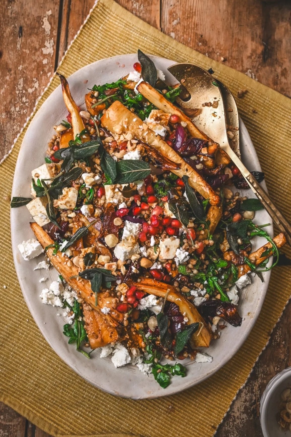 Overhead view of parsnip feta salad on platter with serving spoons, showing layers of buckwheat, roasted vegetables, pomegranate seeds and herbs