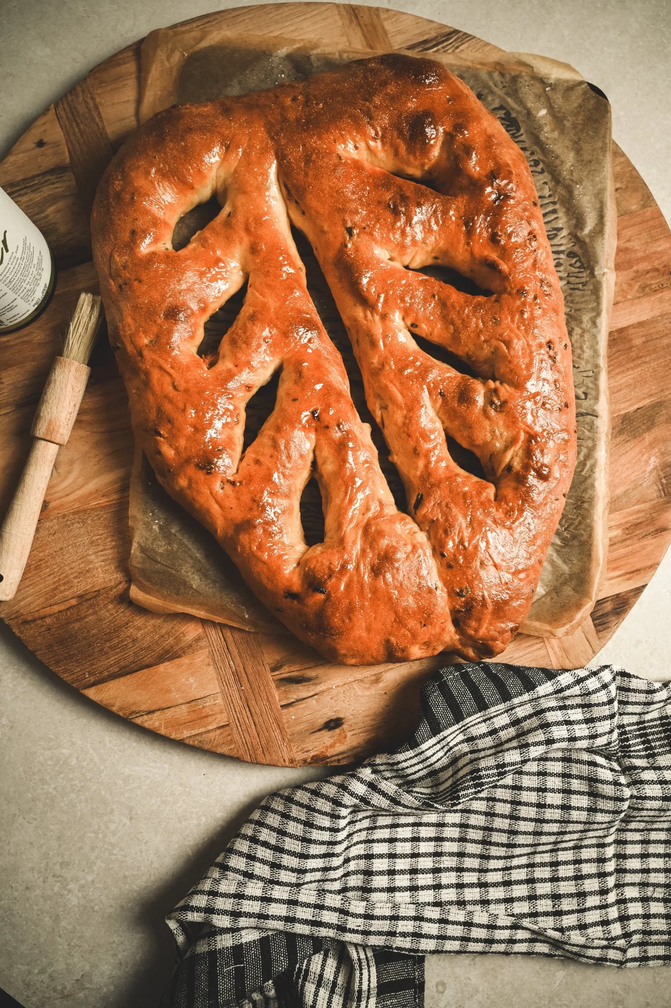 Golden baked sun-dried tomato and oregano fougasse leaf bread on baking paper over a wooden board alongside a pastry brush.