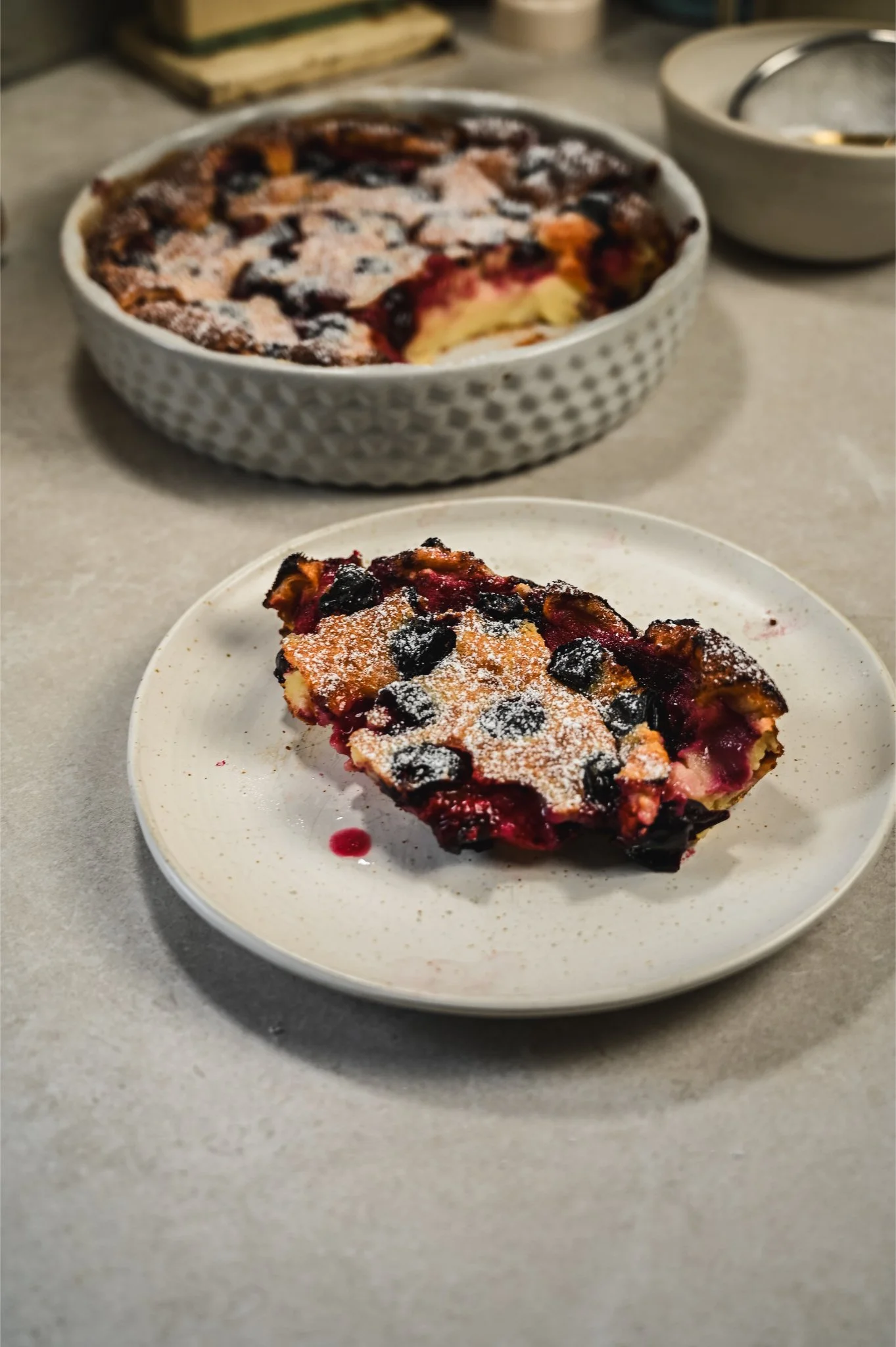 A slice of baked damson and orange clafoutis dusted with icing sugar on a white plate, with the whole ceramic baking dish set in the background.