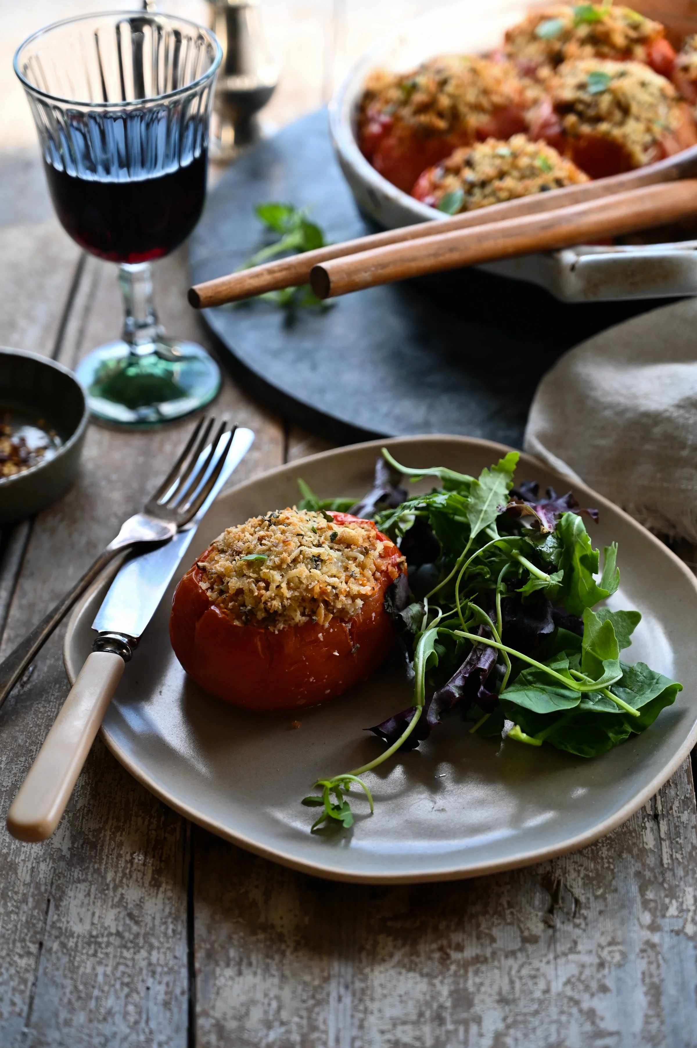 A plated stuffed tomato with salad on a grey plate with vintage cutlery, a glass of red wine, and the full baking dish of tomatoes on a wooden table in the background.