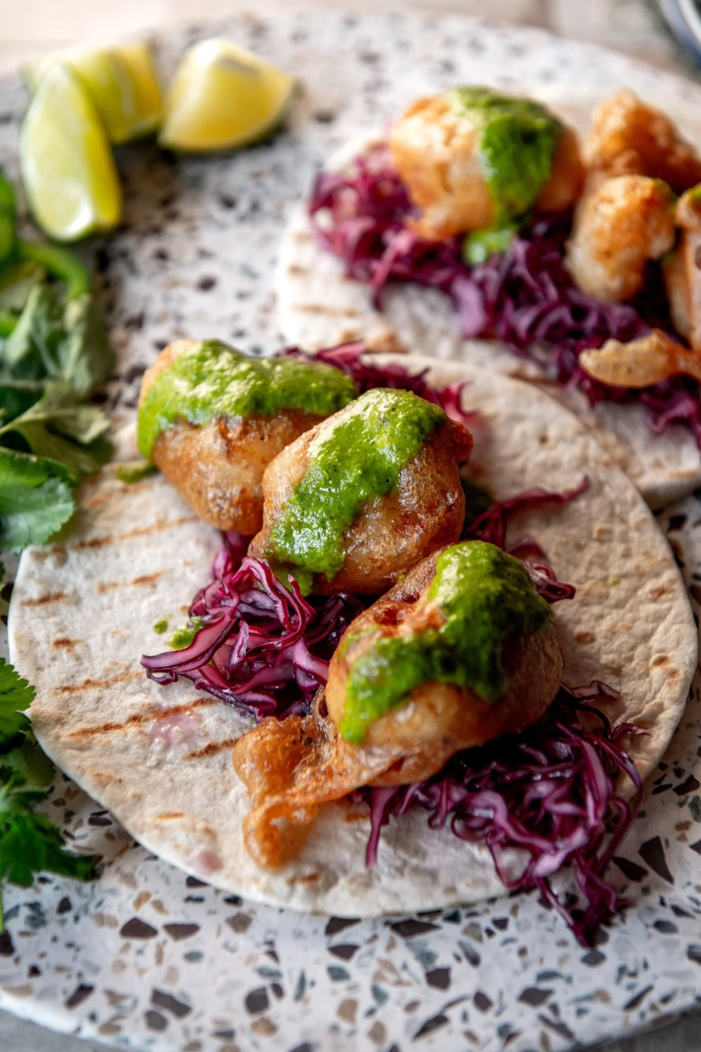 Close-up of a beer battered cod taco showing the lacy, puffed fermented batter and pieces of golden fried cod on a bed of bright pickled red cabbage, finished with a thick drizzle of green jalapeño sauce on a charred flour tortilla.