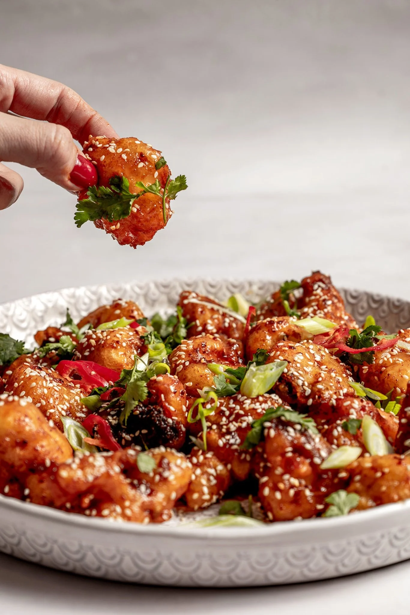 hand holding a single piece of crispy yangnyeom cauliflower coated in glossy gochujang sauce and topped with a leaf of fresh coriander, with the rest of the platter visible below.