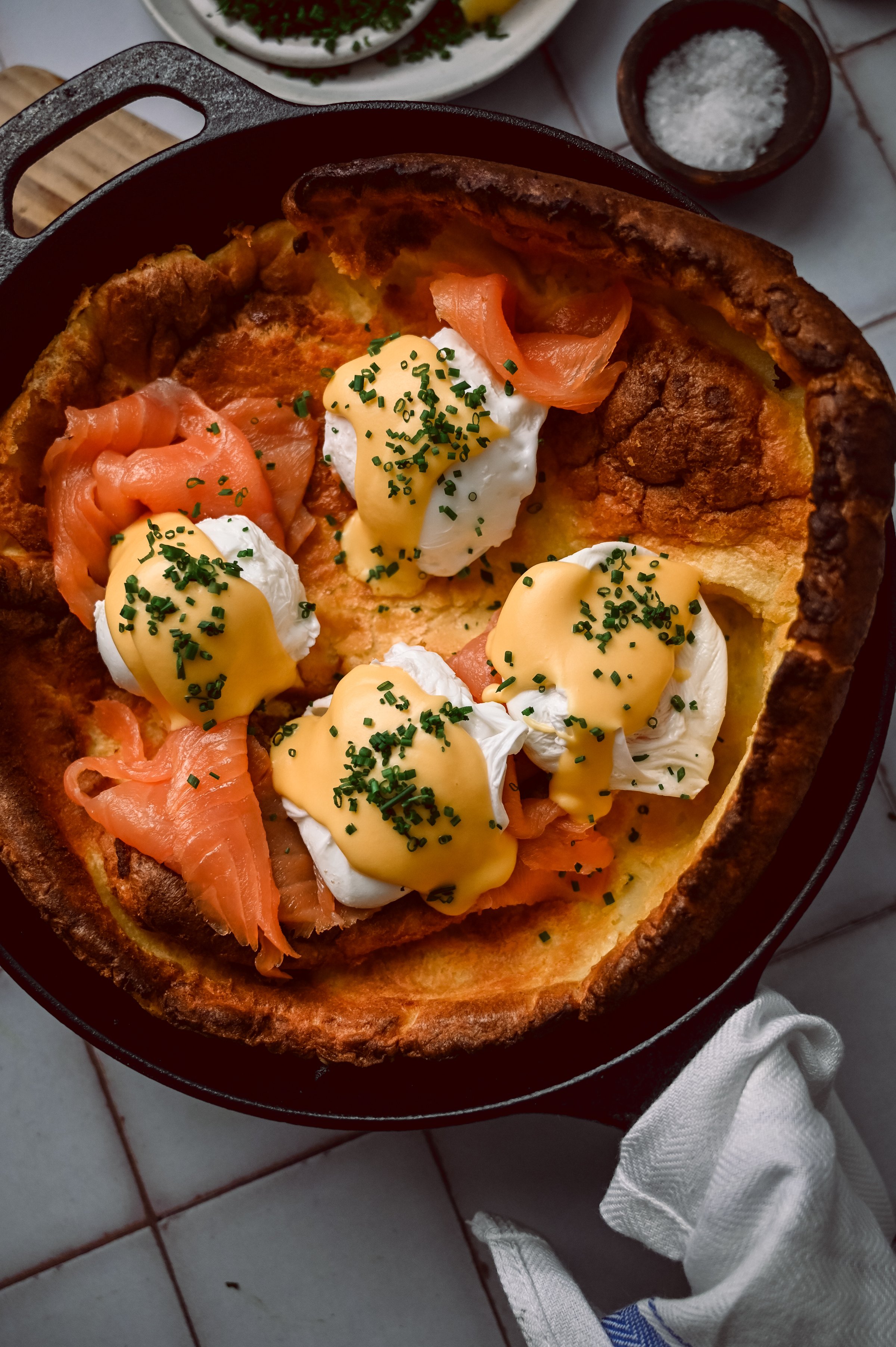 Overhead view of Dutch baby eggs royale in cast iron skillet showing dramatic puffed edges, four poached eggs, layers of smoked salmon, and glossy hollandaise.