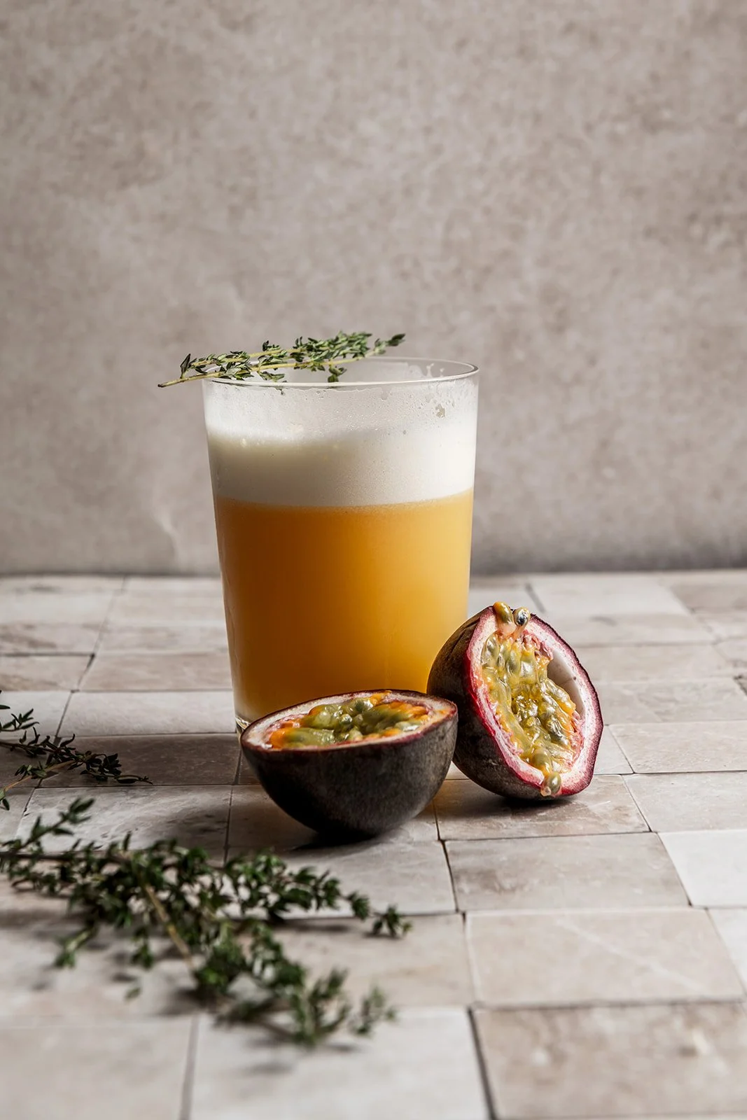 A tall glass of Tropical Thyme Sour on pale stone tiles, with a frothy white top, golden passion fruit juice below, a sprig of fresh thyme resting on the rim and two halved passion fruits beside the glass.