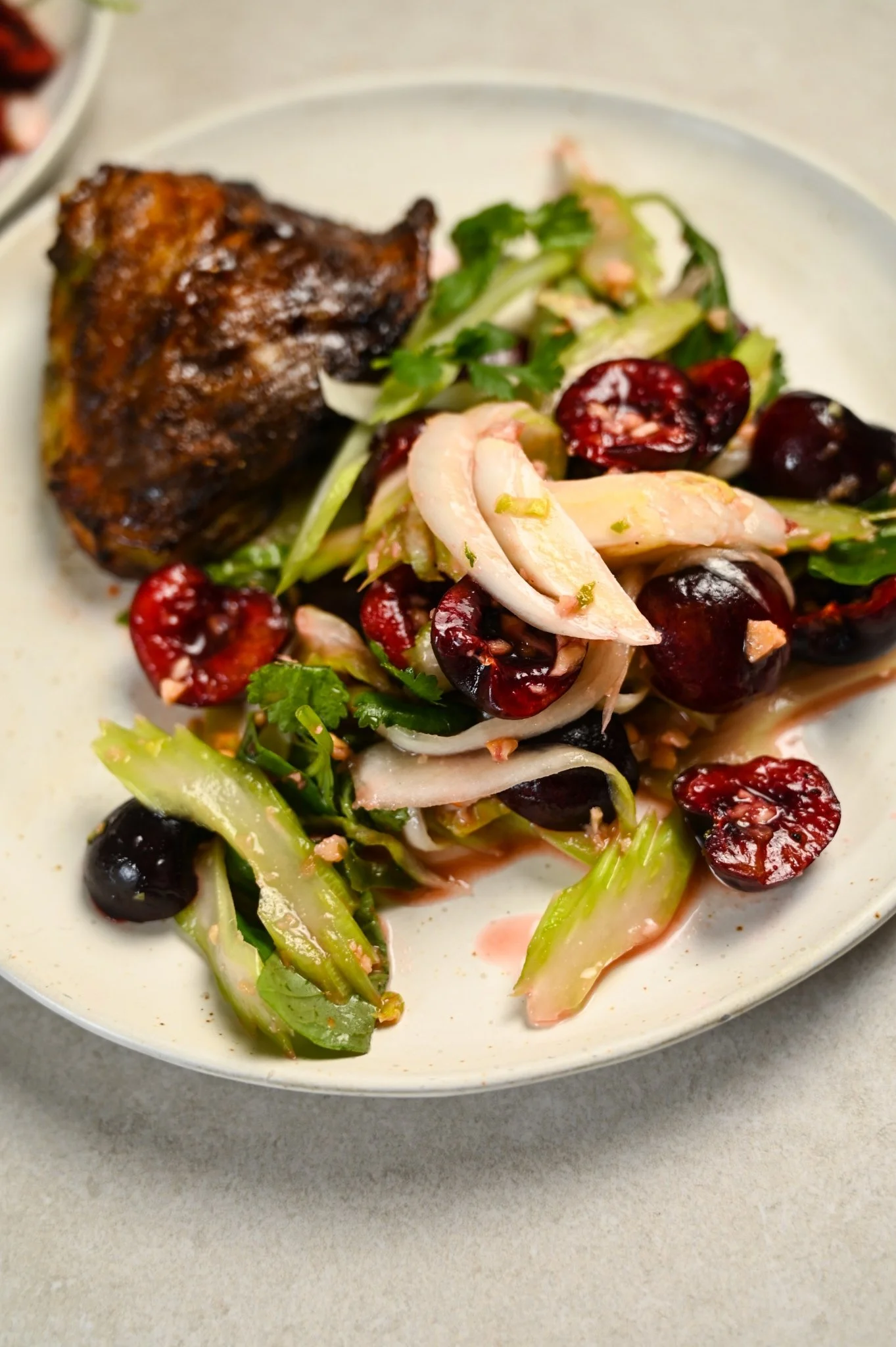 Cherry som tam with halved cherries, sliced celery, shallot and coriander in a glossy dressing, served with a piece of crispy roast chicken on a pale speckled plate.
