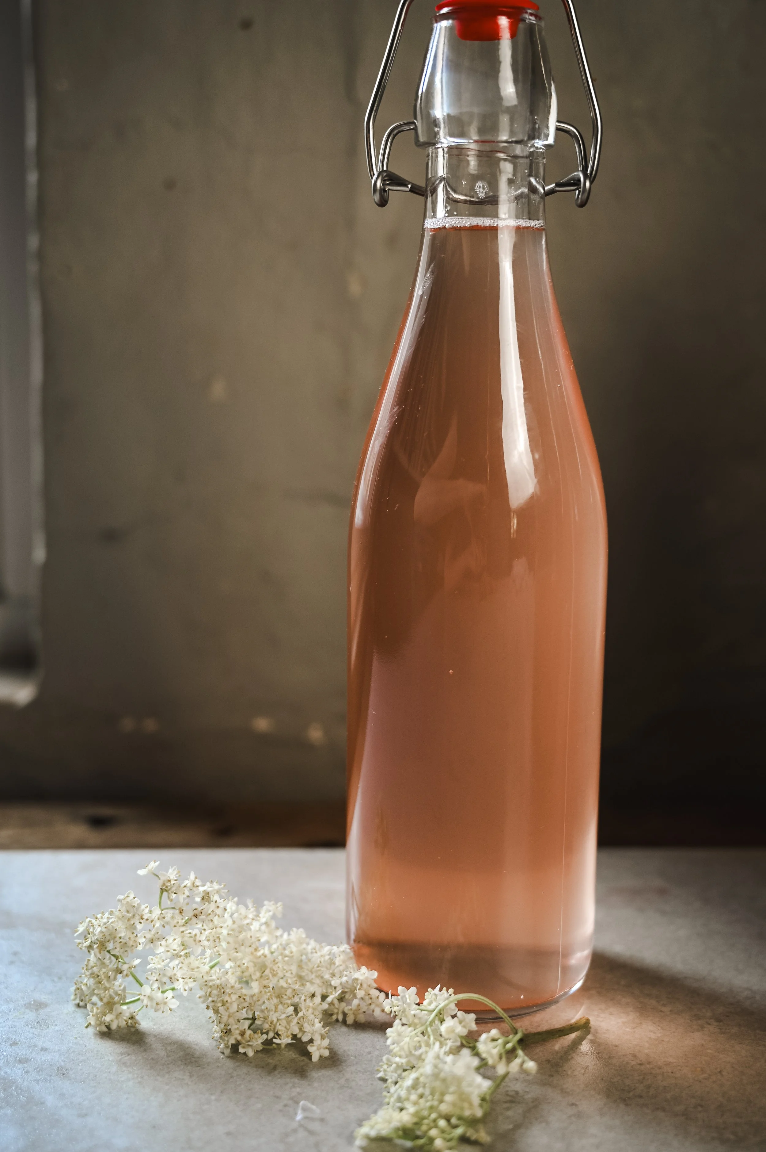 Bottled rhubarb and elderflower cordial, pale pink in a swing-top glass bottle, with fresh elderflower heads laid on a grey stone worktop.