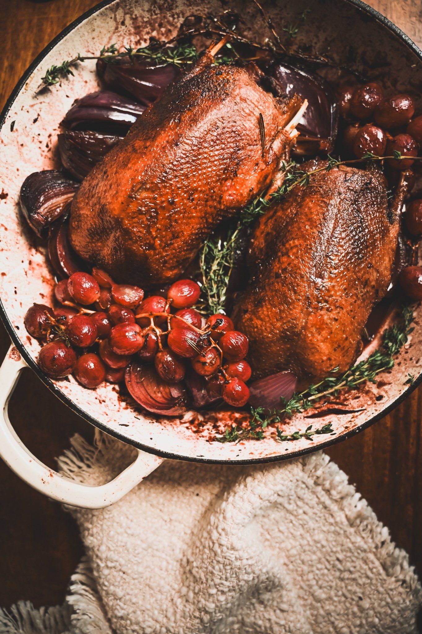 Overhead view of whole roasted wild mallard in Dutch oven surrounded by caramelised red onions and grapes with fresh thyme
