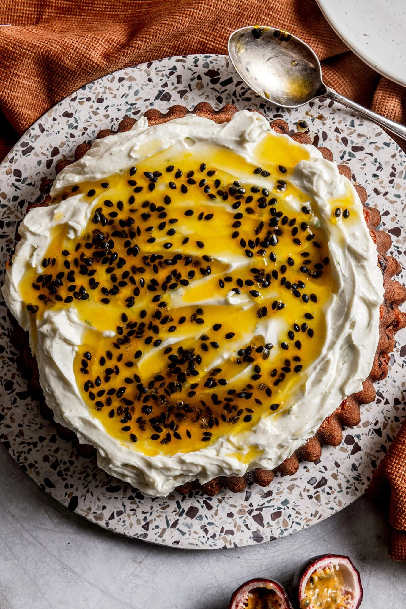 Passion fruit cake with soft mascarpone icing and a seeded passion fruit coulis drizzled over the top, served on a terrazzo plate with a vintage spoon and halved passion fruit alongside.