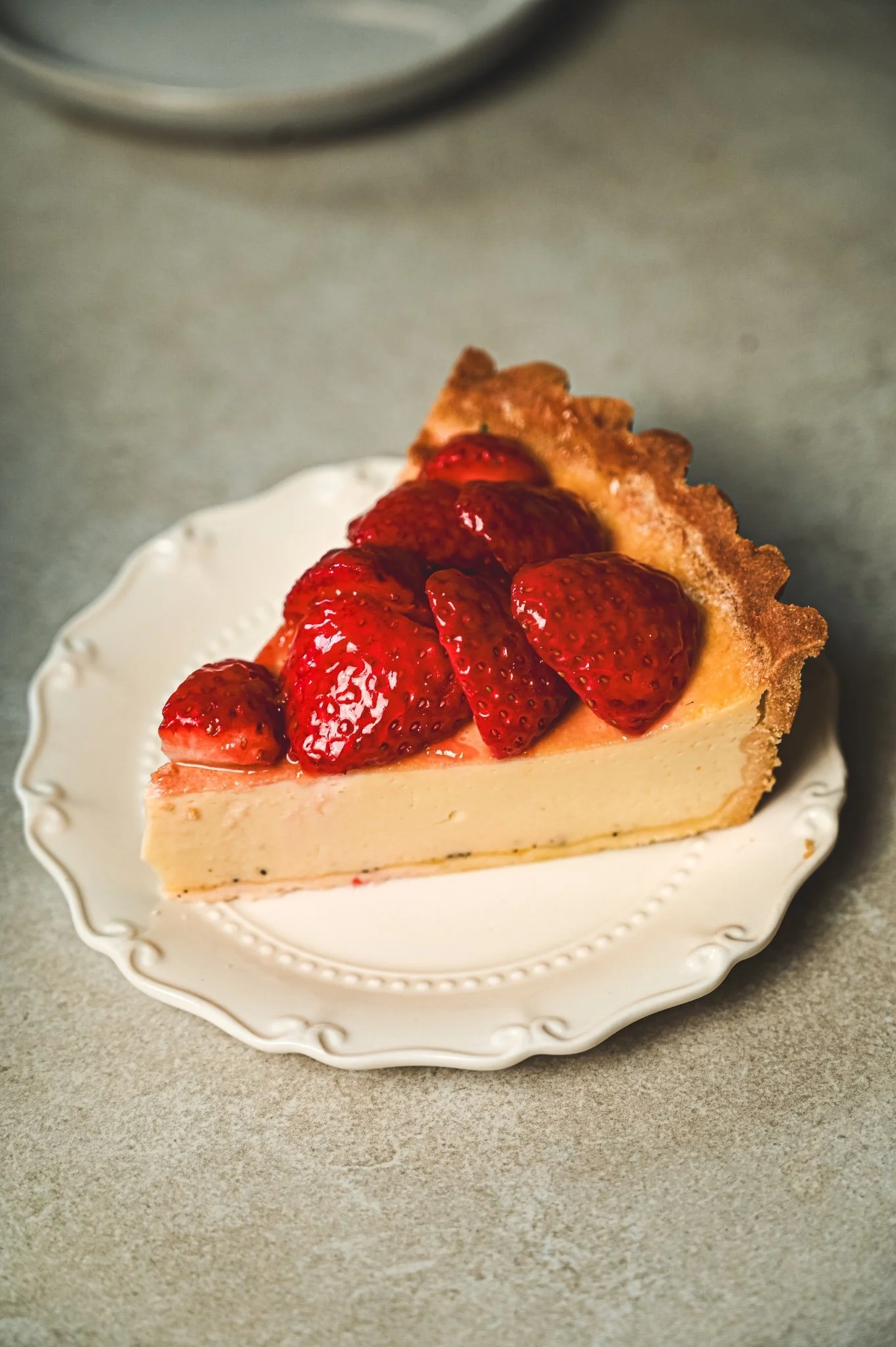 Slice of strawberry tart on a white fluted plate, glazed strawberries over a creamy vanilla-flecked filling in a golden pastry crust.