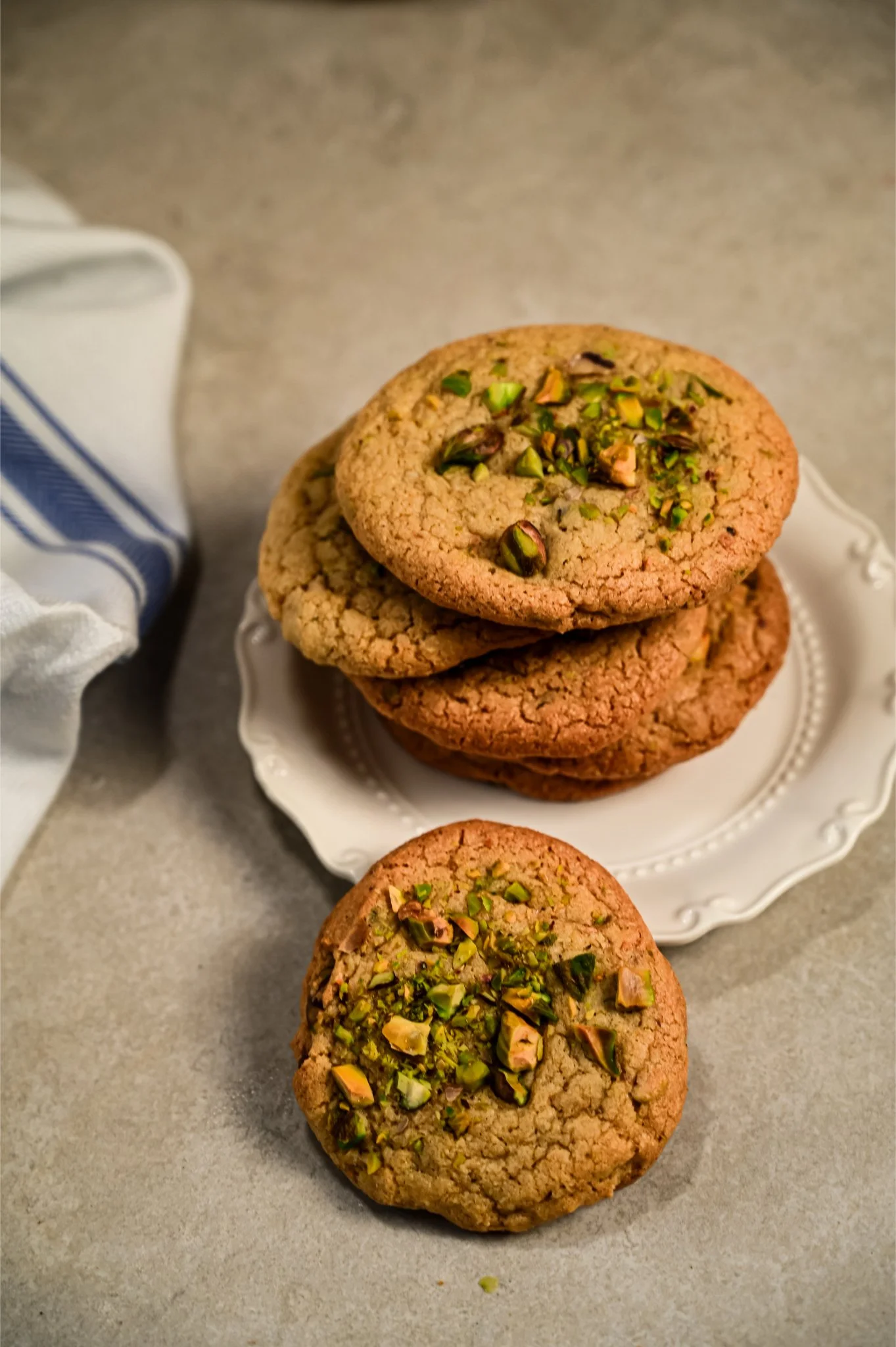 Stack of golden Persian love cookies topped with bright green chopped pistachios on a scalloped white plate, alongside a blue-striped tea towel.