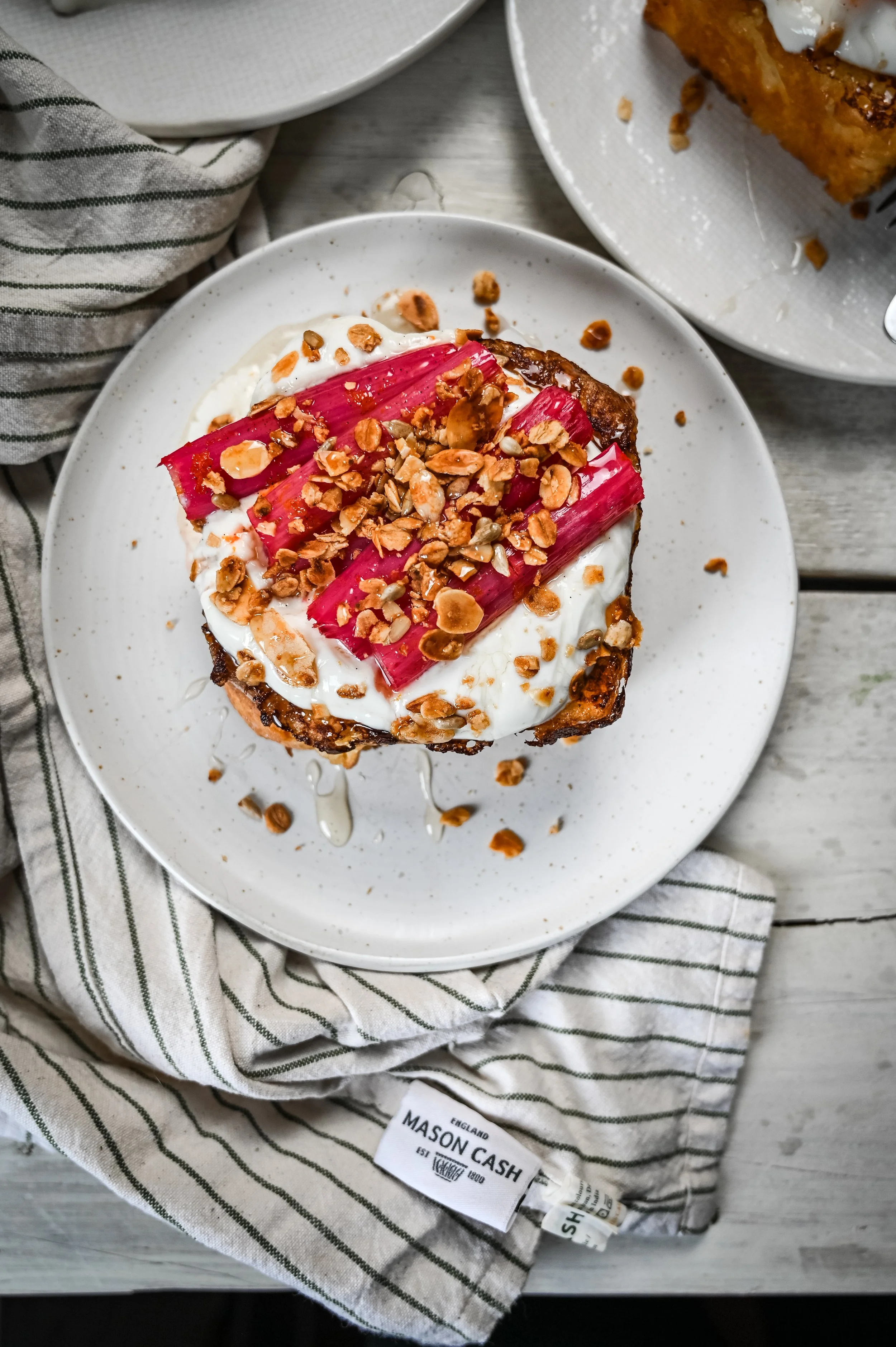Overhead shot of brioche French toast topped with roasted rhubarb and ginger granola on a white speckled plate, styled with a striped linen cloth
