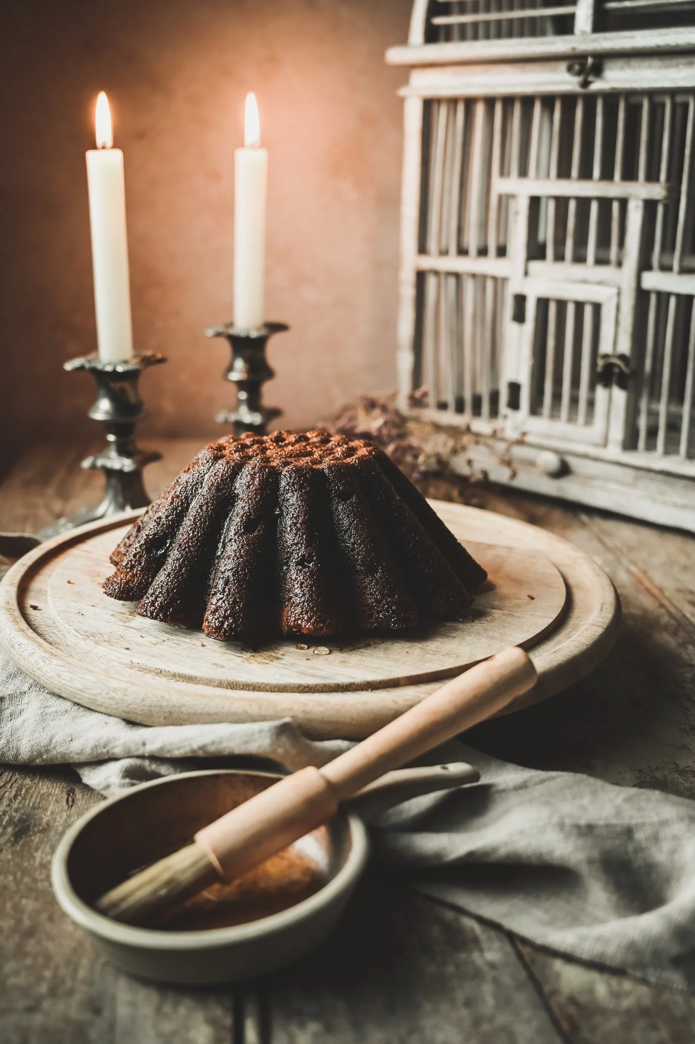 Gluten-free ginger and sultana Bundt cake being brushed with glossy ginger syrup on a rustic wooden board beside lit candles.