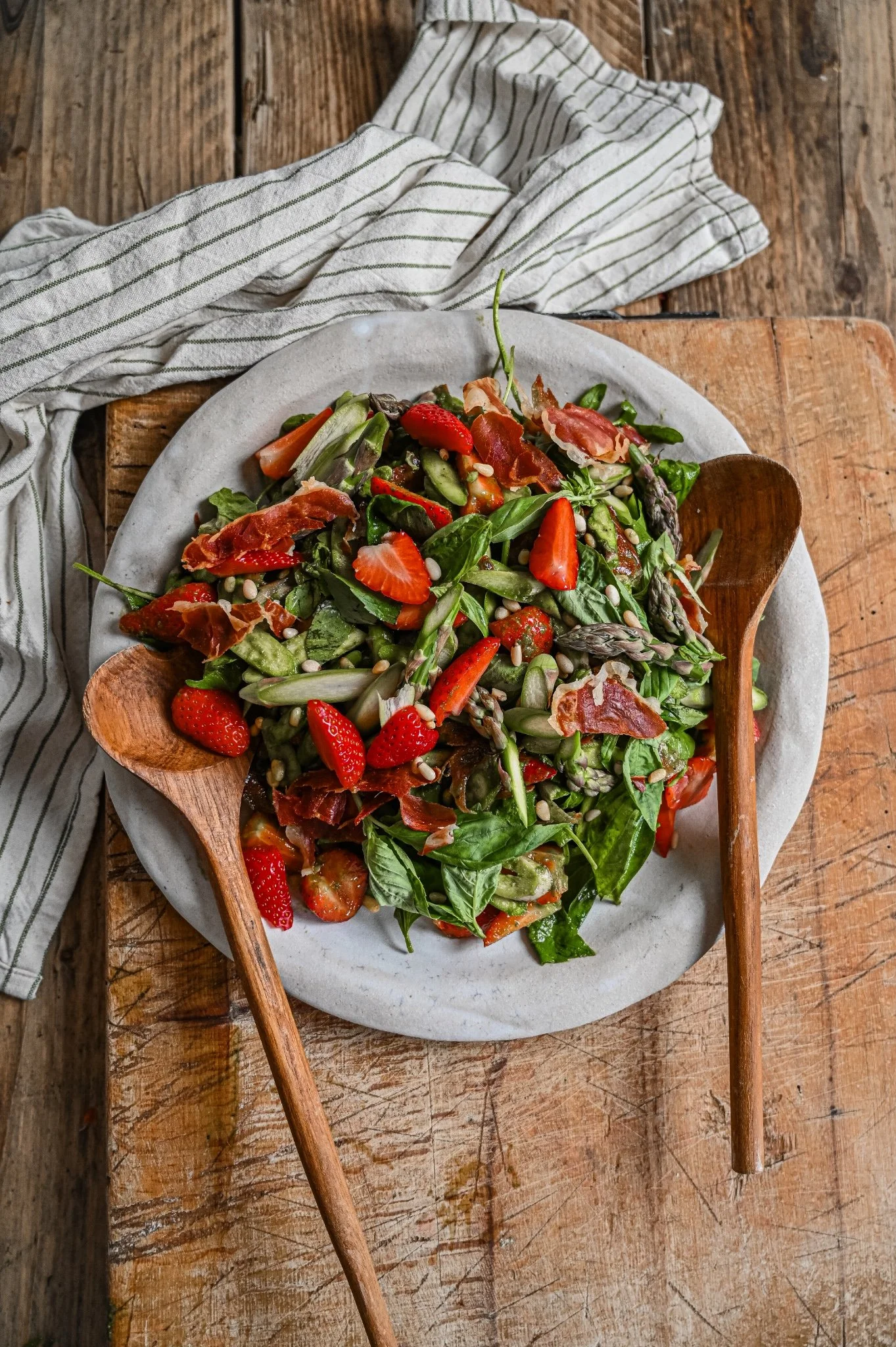 Strawberry and asparagus salad with crispy Serrano ham, fresh basil and toasted pine nuts, served on a stoneware plate with two wooden serving spoons on a rustic wood board.