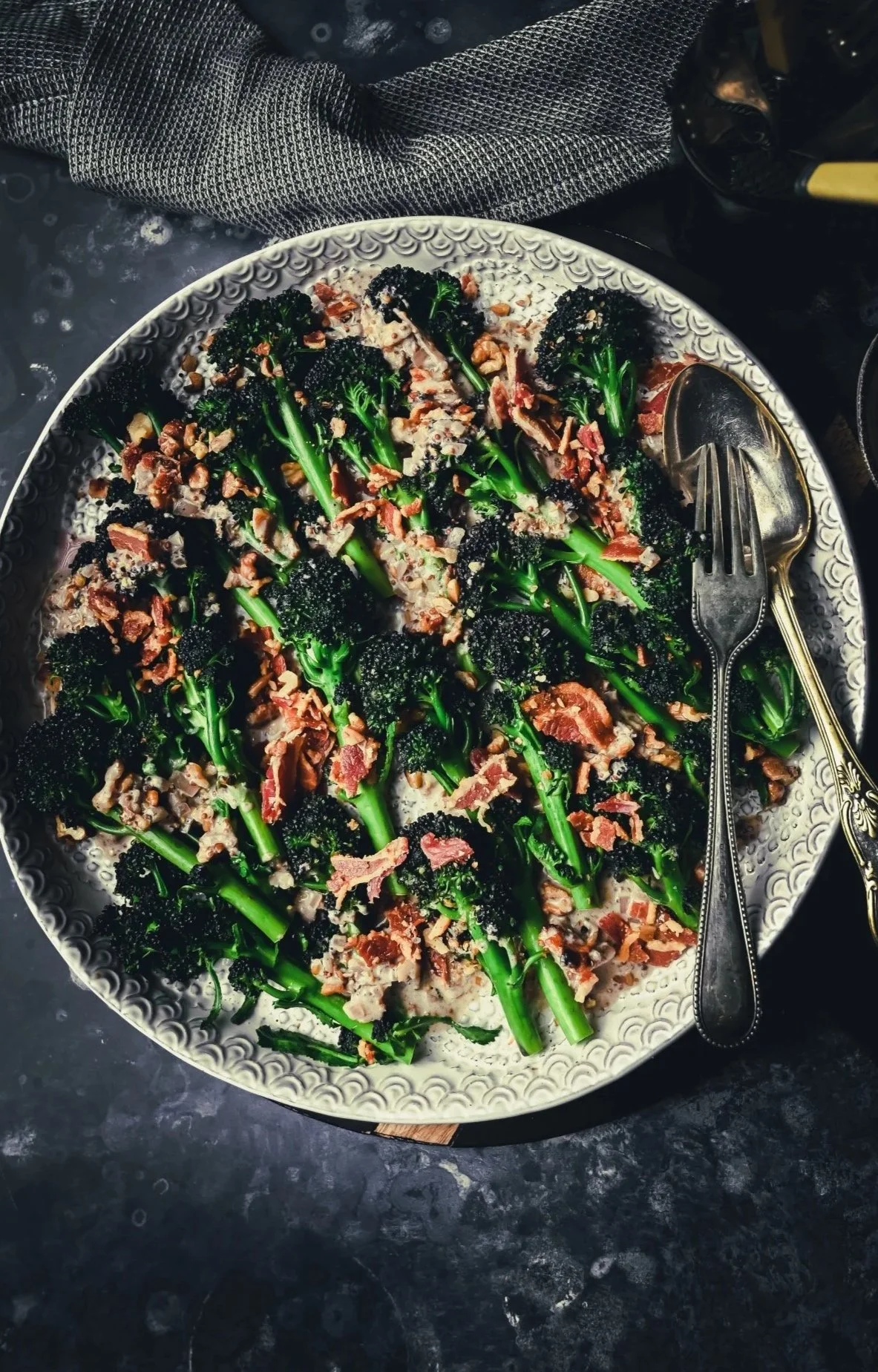 Purple sprouting broccoli with crispy bacon shards and toasted walnuts on white platter with creamy mustard sauce