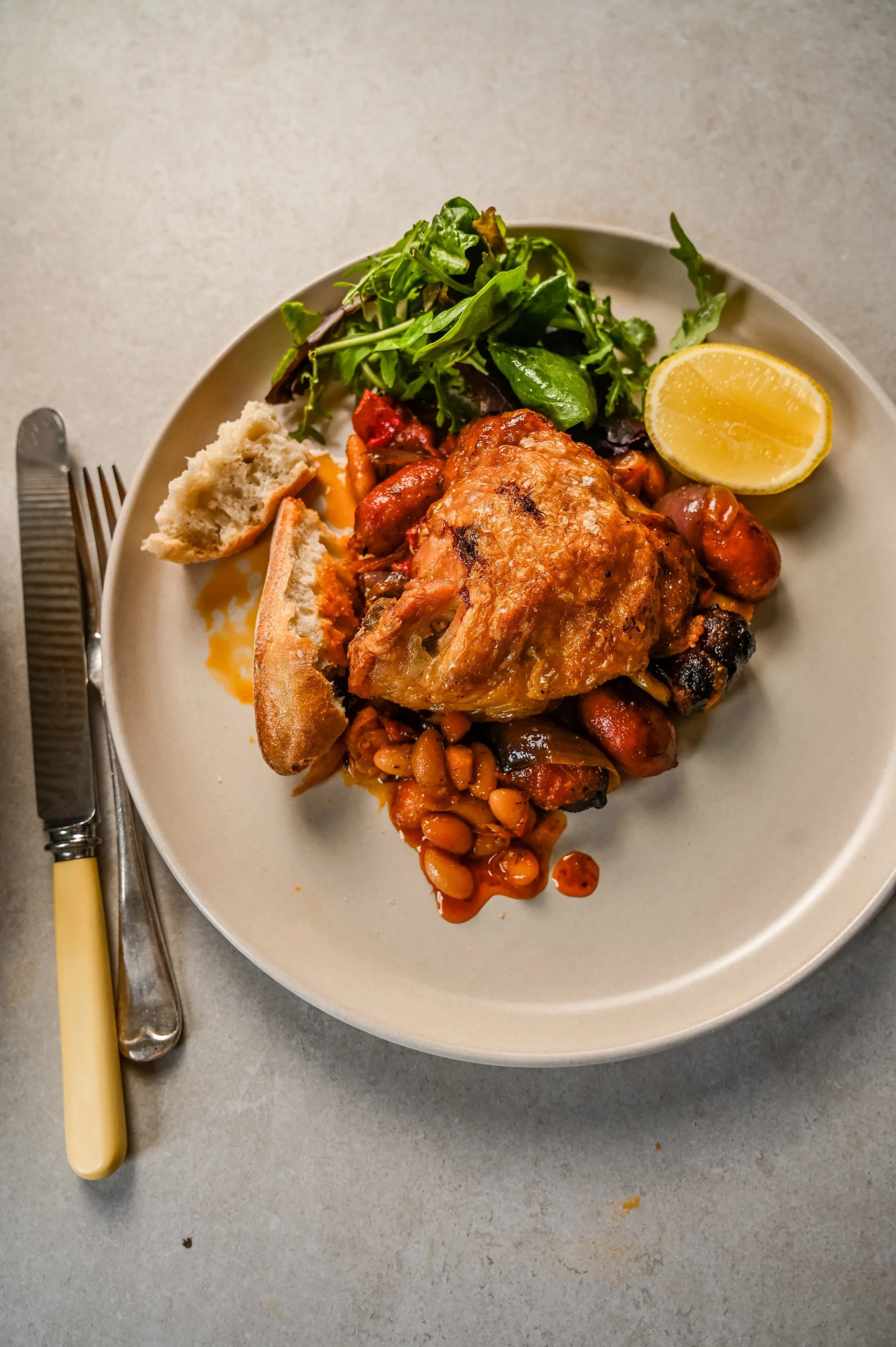 Plated serving of a crispy chicken thigh resting on butter beans, mini cooking chorizo, roasted peppers, and red onions, served alongside fresh green salad leaves, a lemon half, and crusty bread.