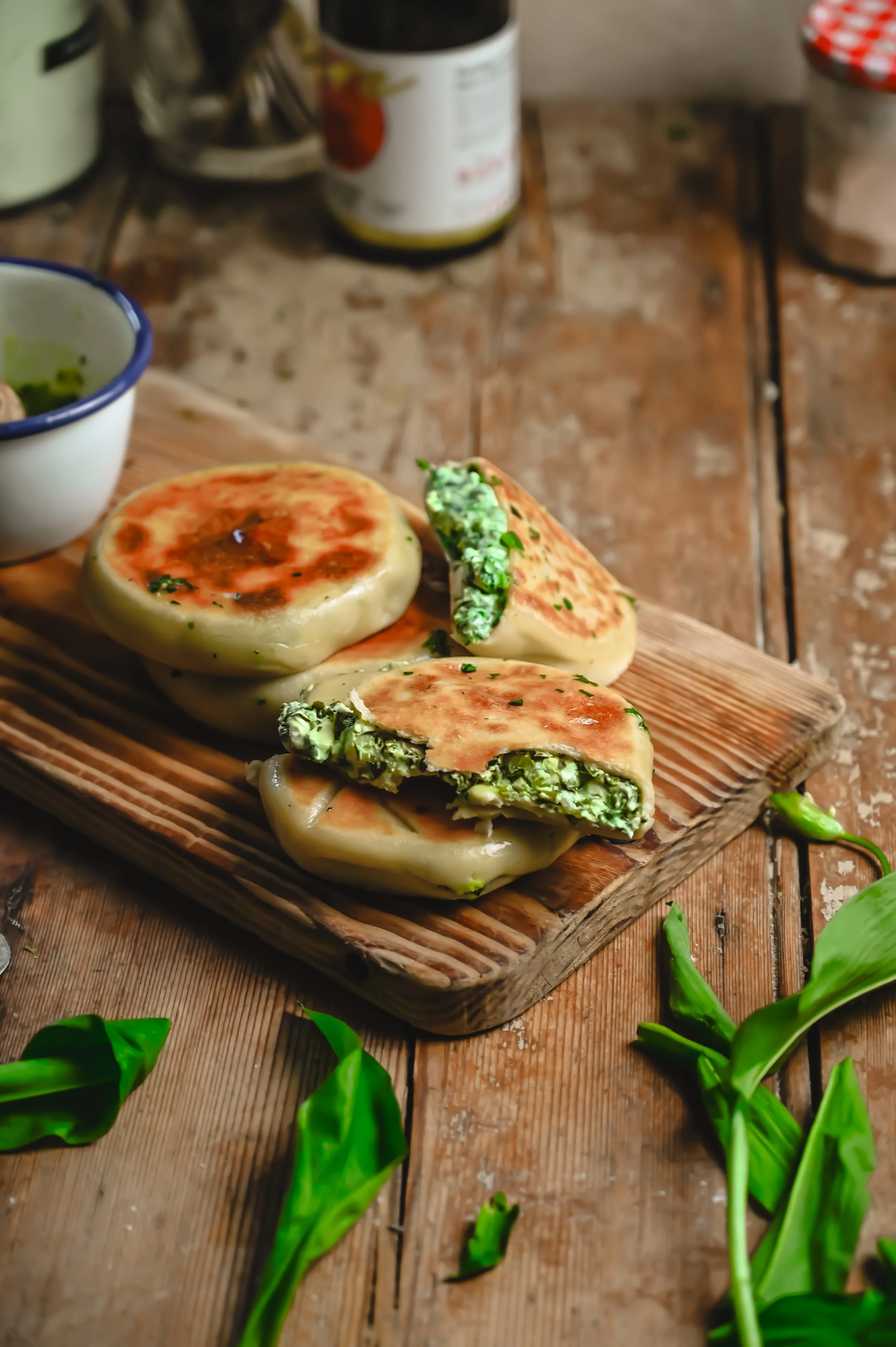 Wild garlic and feta flatbreads on a wooden board, one split open to show the generous green filling, with fresh wild garlic leaves scattered around