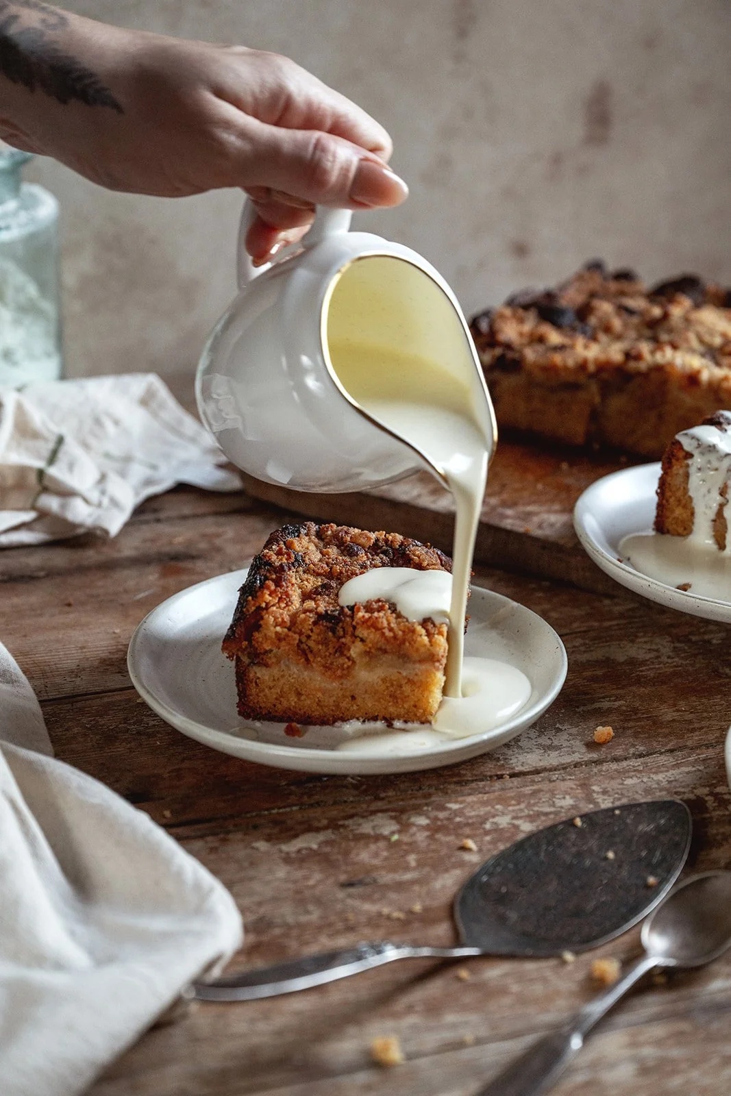 Slice of apple crumble cake on white plate showing layers of sponge, caramelized apples and golden crumble topping with a jug of cream being poured over.