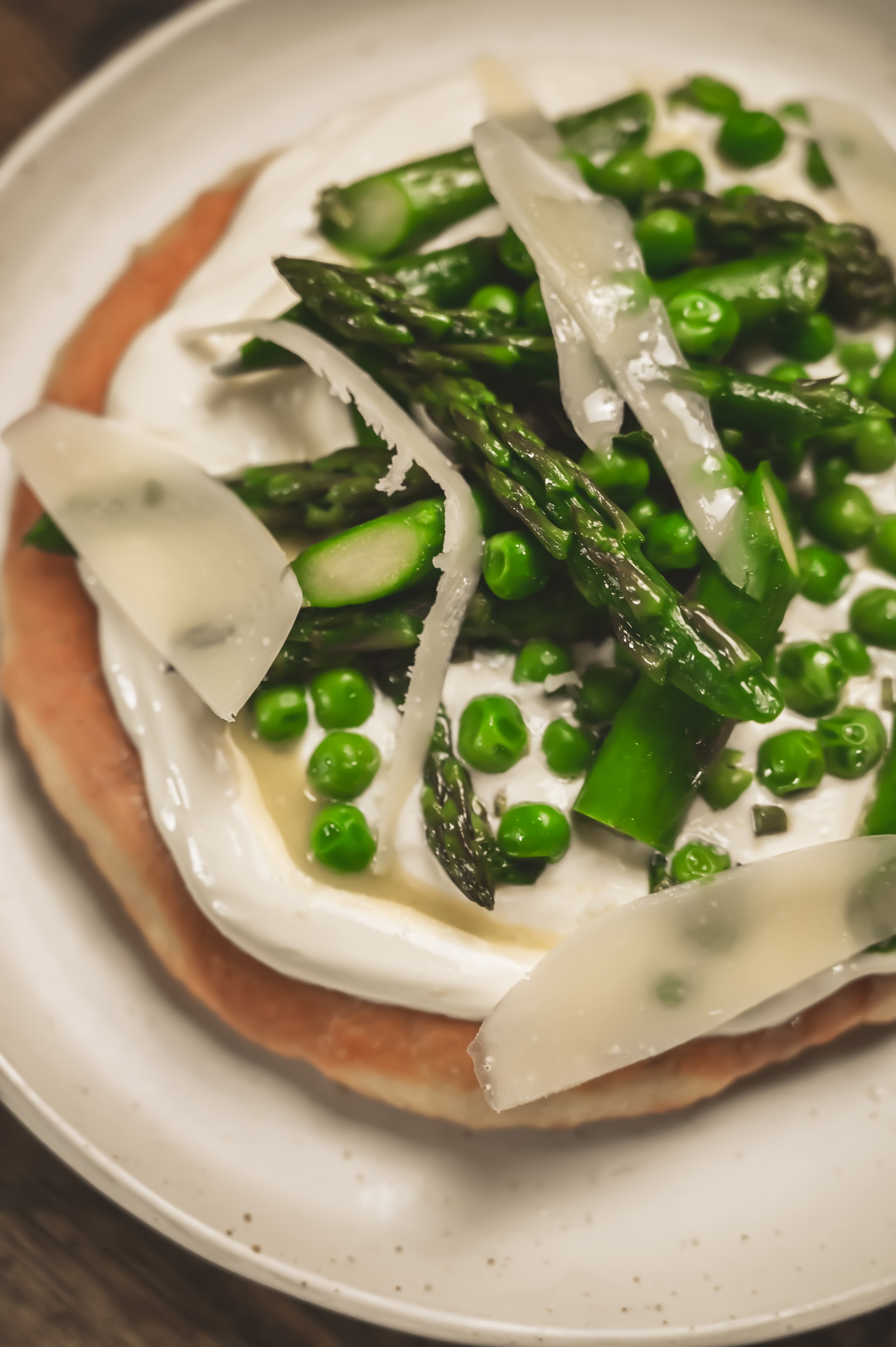 overhead, close crop, lots of pecorino, slightly darker tone) Close overhead shot of a spring lángos with asparagus, peas and generous pecorino shavings over sour cream on golden fried dough