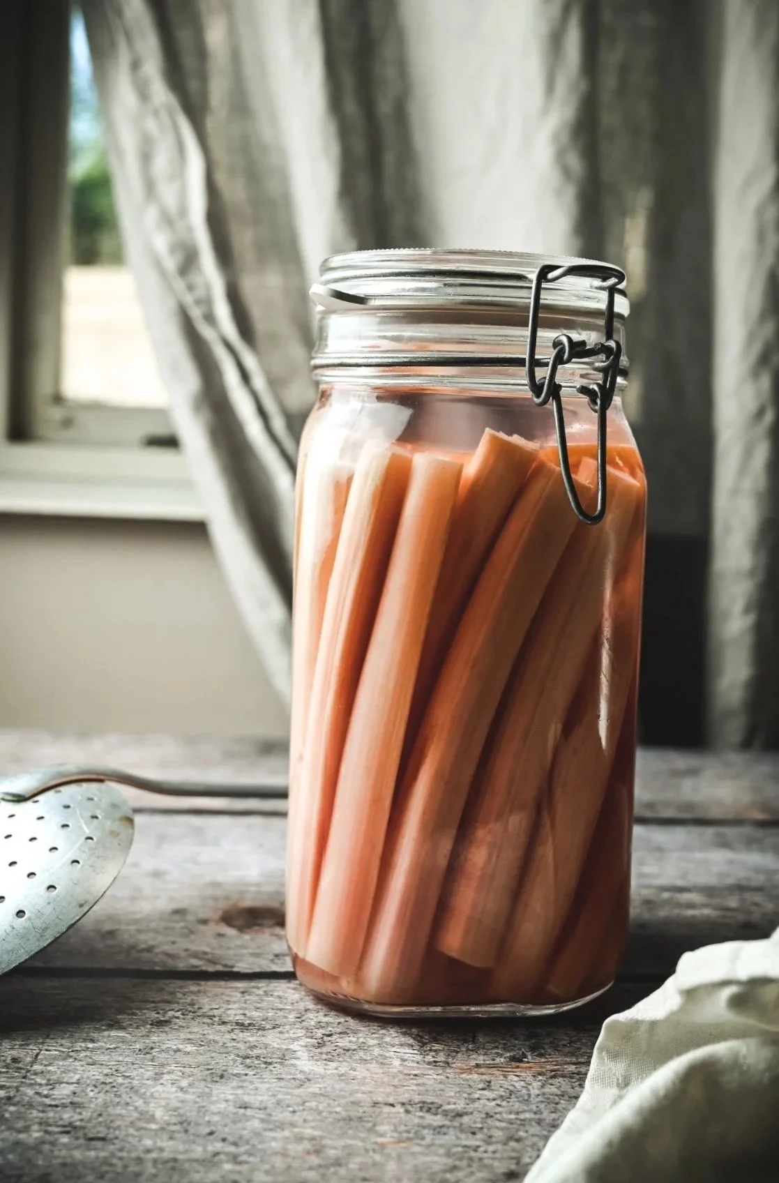 Fermented rhubarb stalks packed upright in a glass clip-top jar on a rustic wooden surface with linen cloth and vintage strainer, natural window light