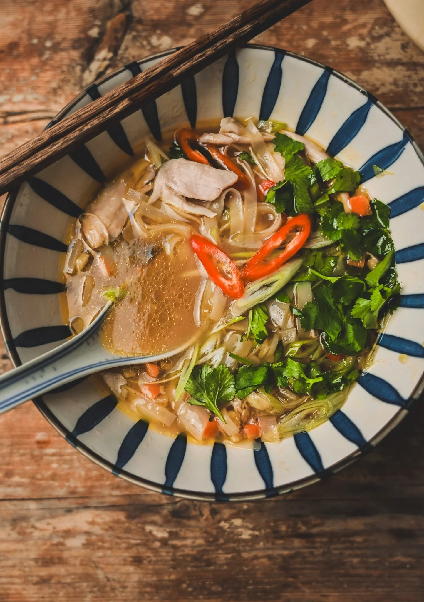 Bowl of Asian chicken noodle soup with thick rice noodles, shredded chicken, fresh coriander and lime wedge on rustic wooden table