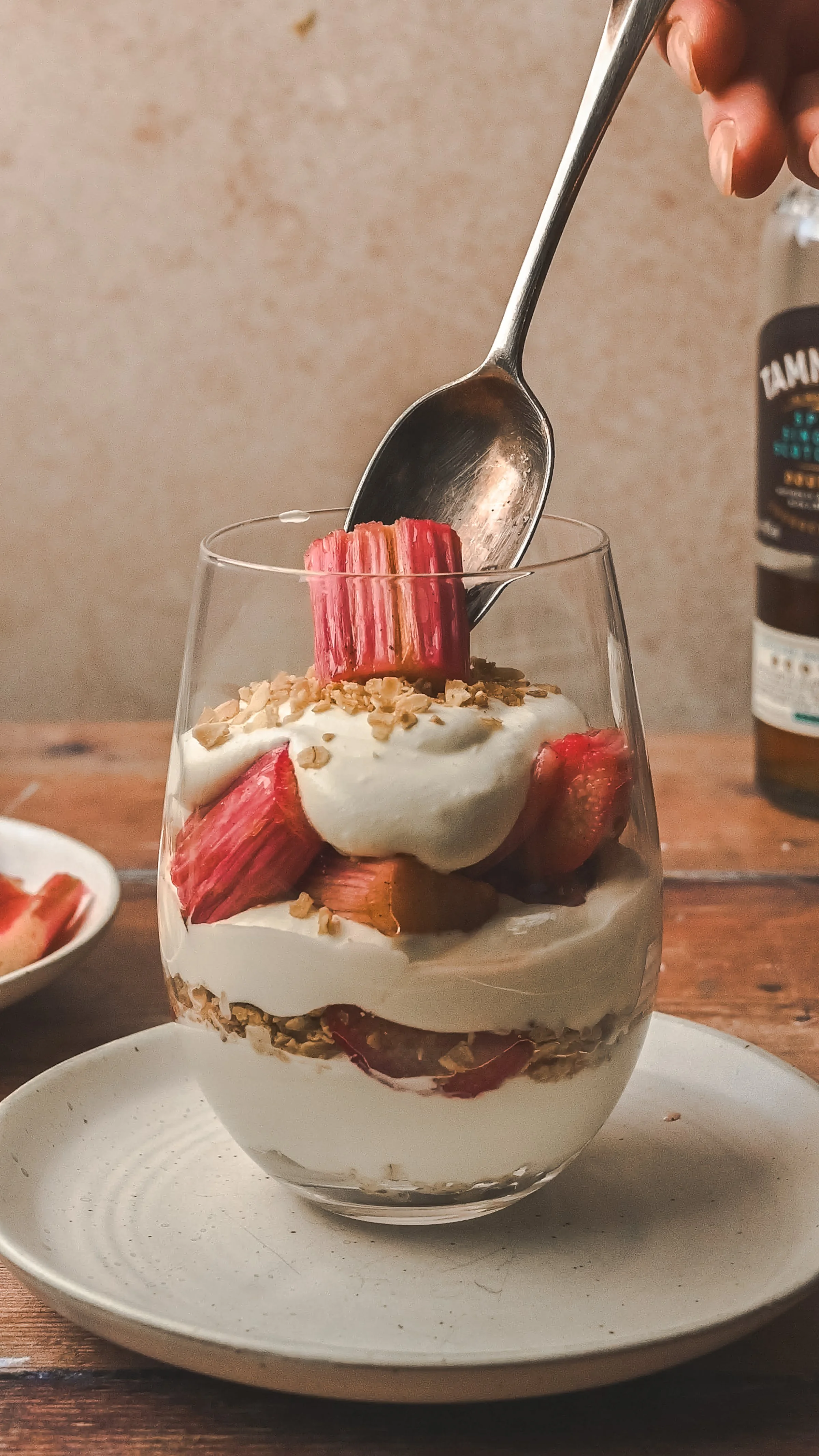 Single glass of rhubarb cranachan on a white plate with a bottle of Scotch whisky and fresh rhubarb stalks visible in the background