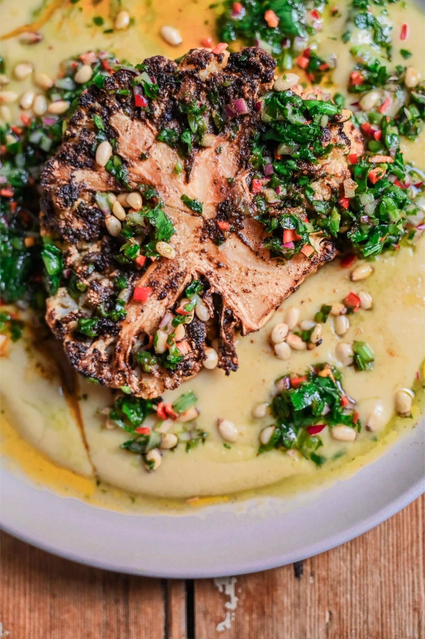 Close overhead shot of a spice-rubbed cauliflower steak on white bean purée, generously dressed with wild garlic chimichurri, pine nuts and olive oil pooling on a grey plate