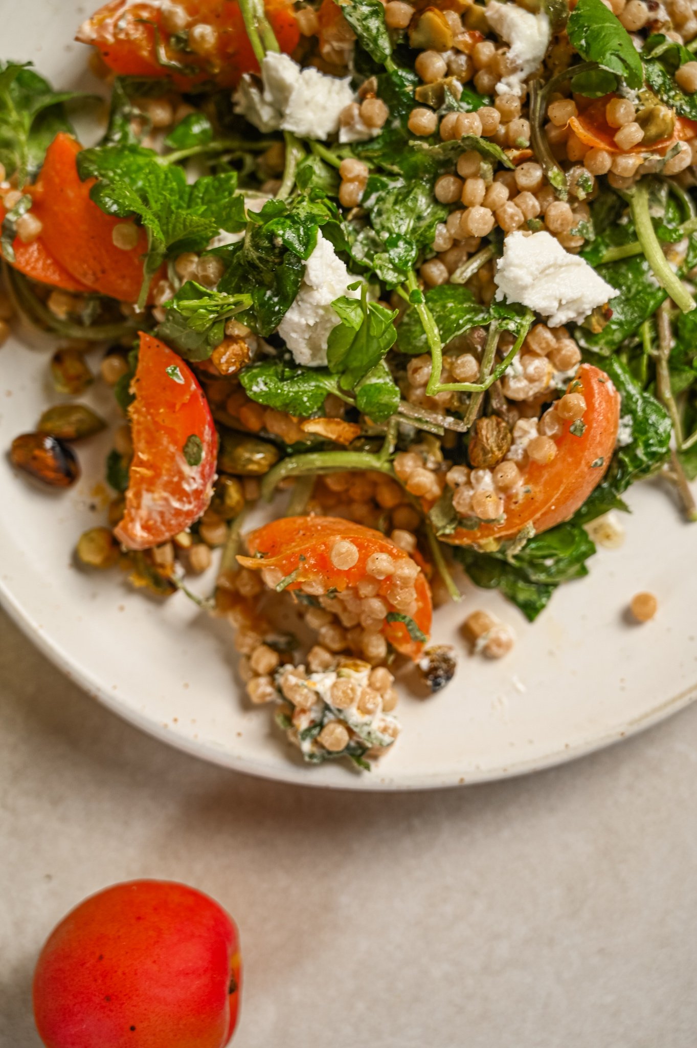 Apricot and fregola salad with roasted apricots, crumbled goat's cheese, watercress, mint and pistachios on a pale speckled plate.
