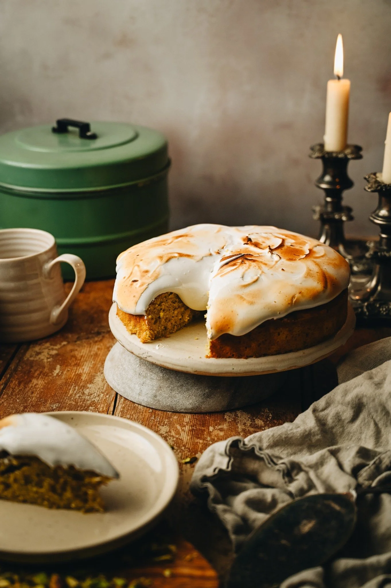 Lemon and pistachio cake on white cake stand with tall torched Italian meringue topping showing golden peaks, lit candles and tea cup in background