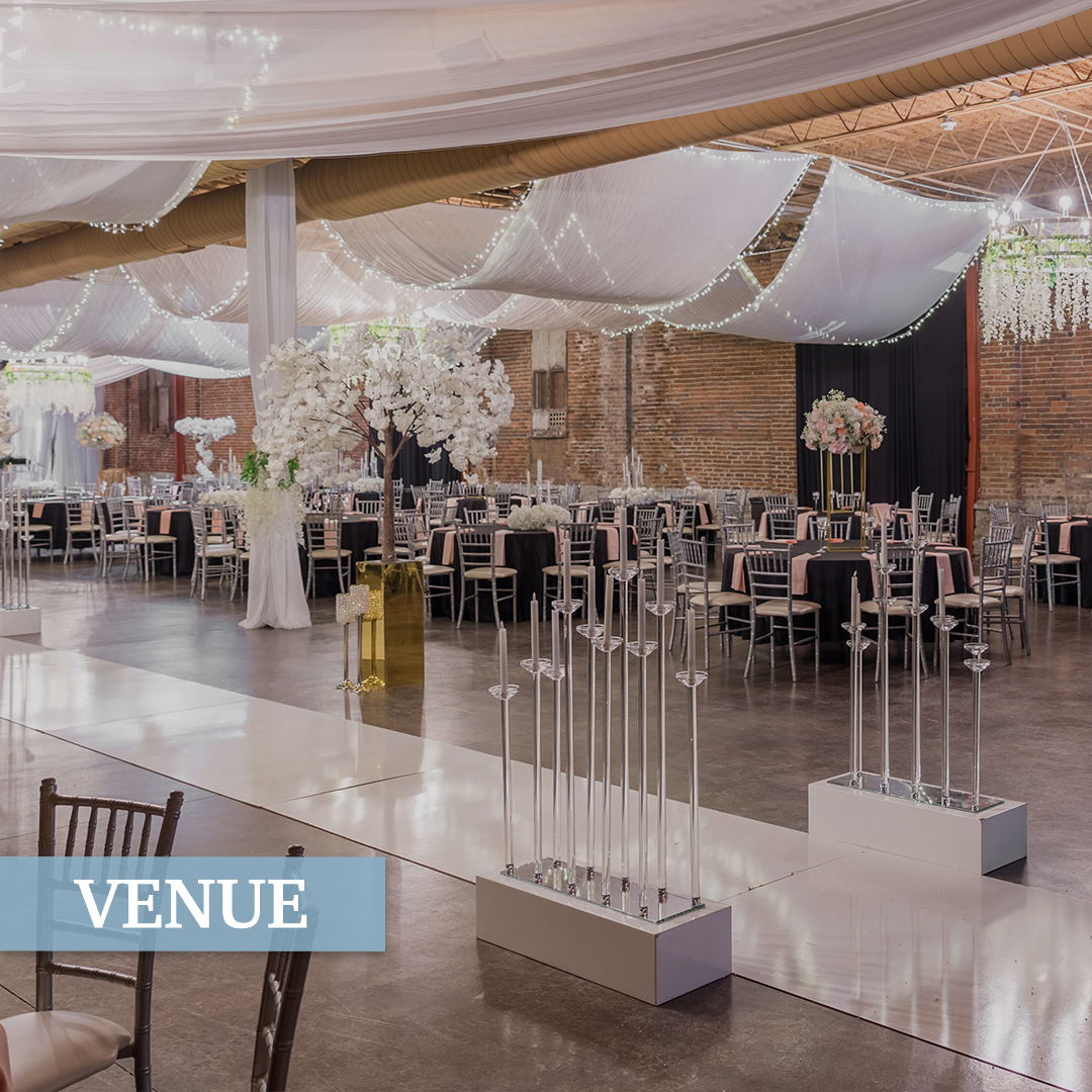 Elegant event venue decorated with white drapes, string lights, and floral centerpieces on round tables with black tablecloths, silver chairs, and tall candle holders.