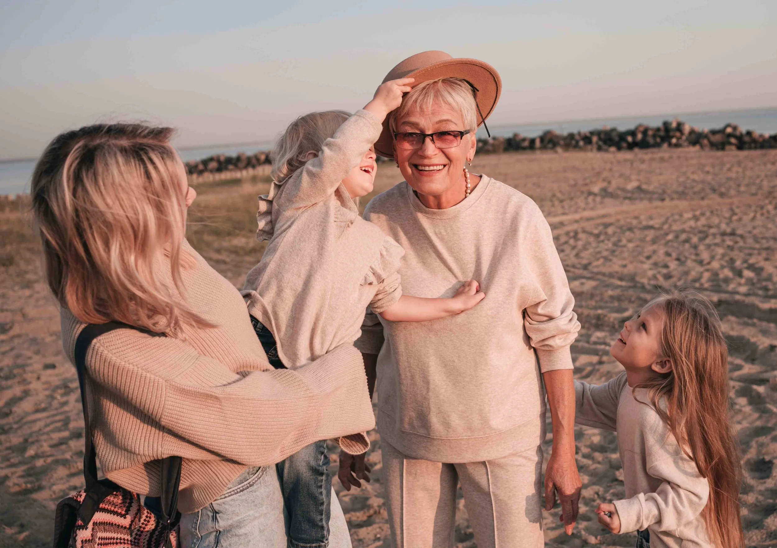 A smiling elderly woman in a beige sweater and glasses wearing a straw hat, standing on a beach with four young girls. One girl is putting the hat on her, others are looking at her and smiling, all dressed casually, with the ocean and rocks in the background at sunset.