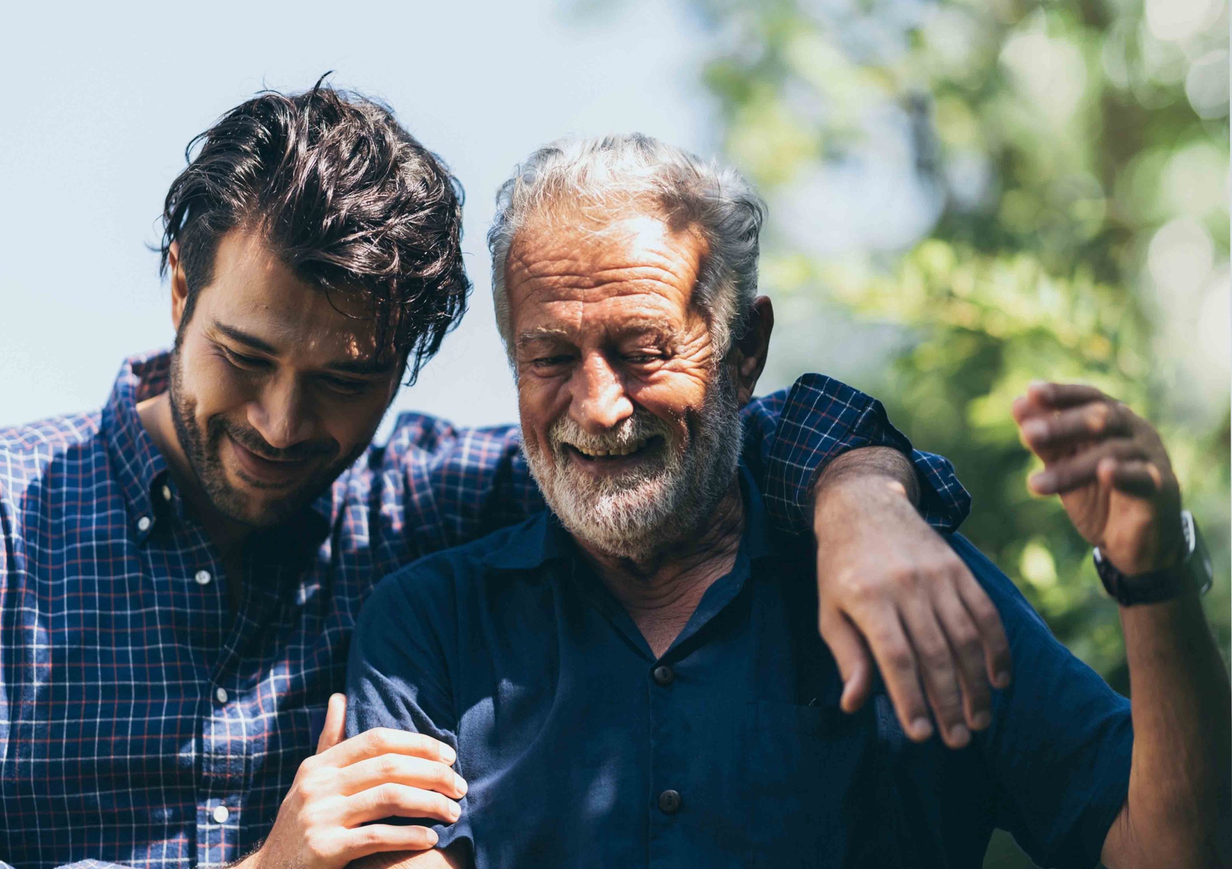 A young man and an older man smiling and celebrating together outdoors, with the young man resting his arm on the older man's shoulder.