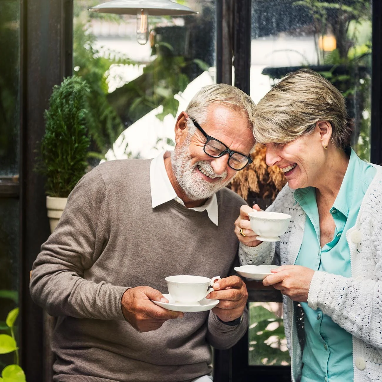 An elderly man and woman sharing a moment, laughing and enjoying tea together in a cozy indoor setting with lush greenery outside.