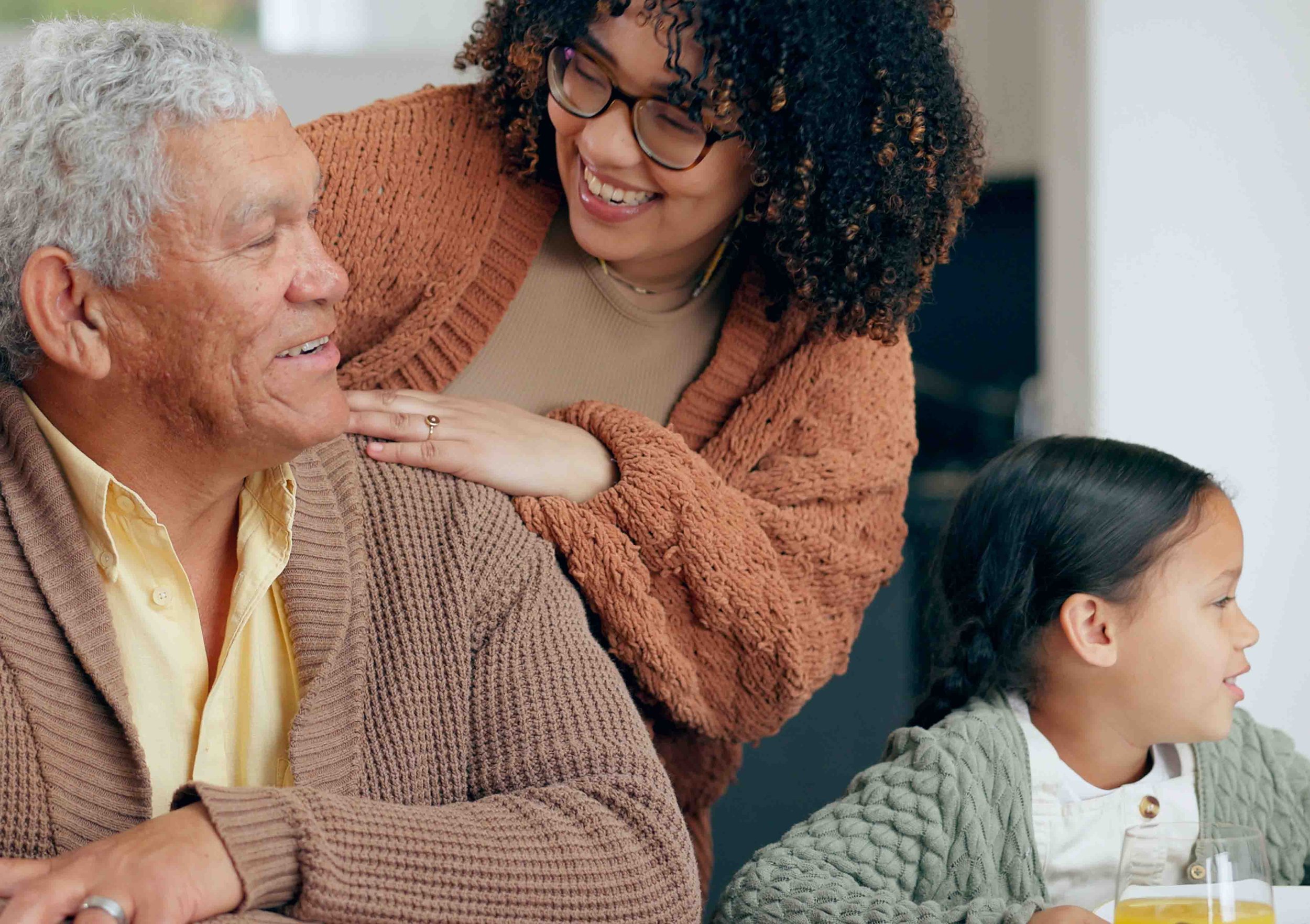 An elderly man with gray hair smiling as a young woman with glasses and curly hair embraces him. A young girl with braided hair sits at the table, looking to the side with a glass of orange juice in front of her.