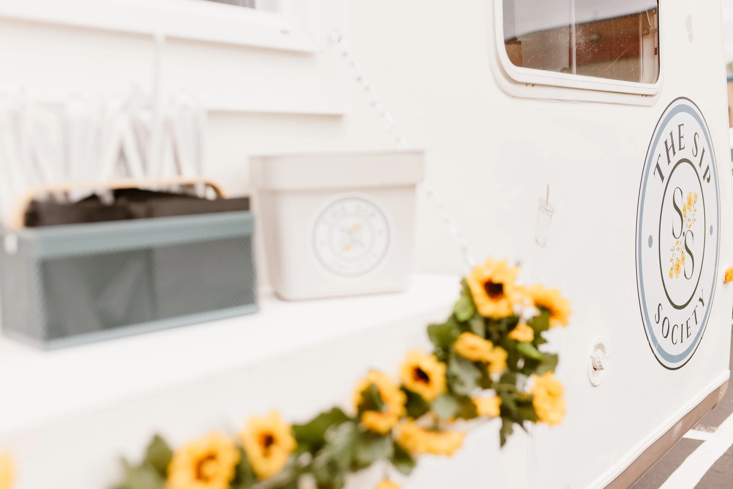 Close-up of a white food truck with a circular logo and yellow flowers in the foreground.
